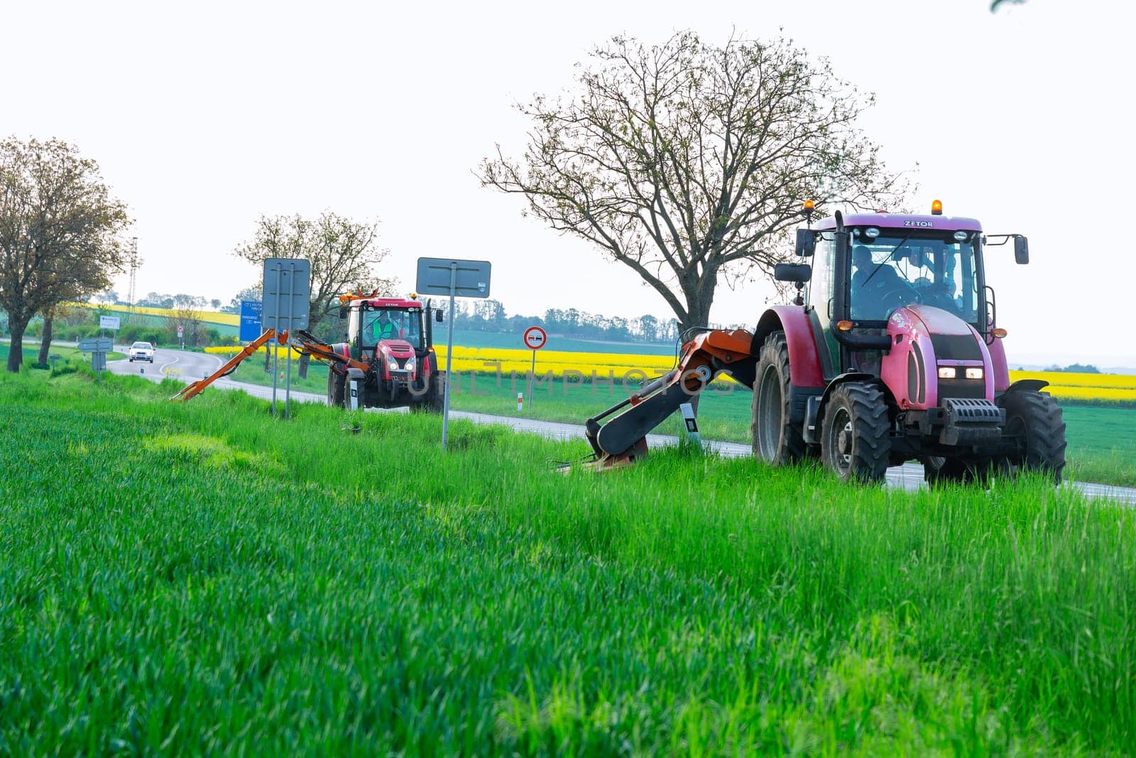 11 May 2022 Crudim, Czech Republic. Rotary mower attached to a tractor moving along the side of the road is mowing the green grass next to the roadway. Clean roadside.