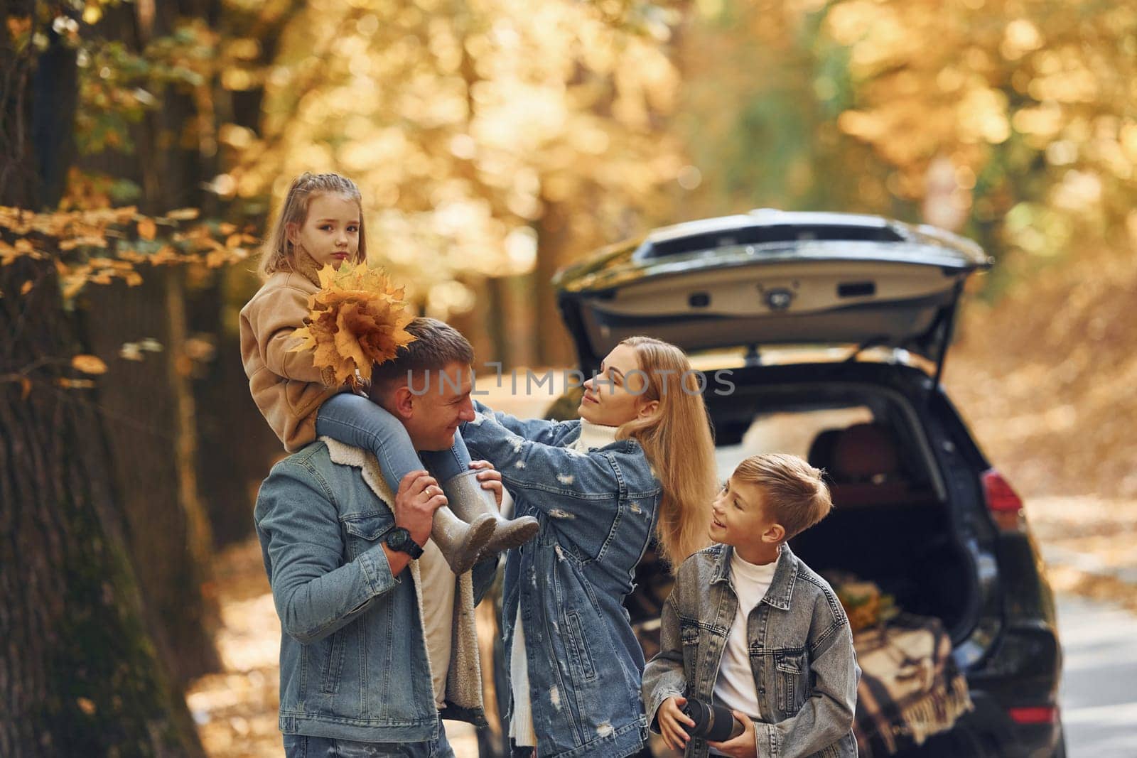 Near car with open trunk. Happy family is in the park at autumn time together.