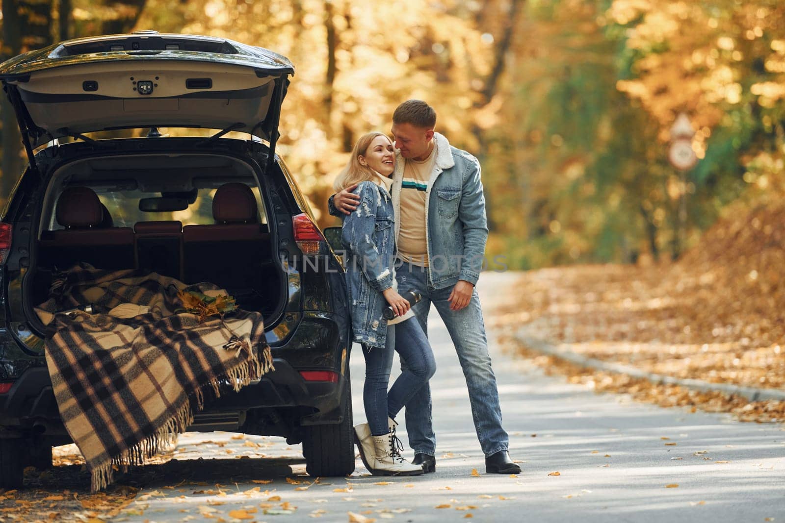 Opened trunk. Couple standing on the road in park near automobile.