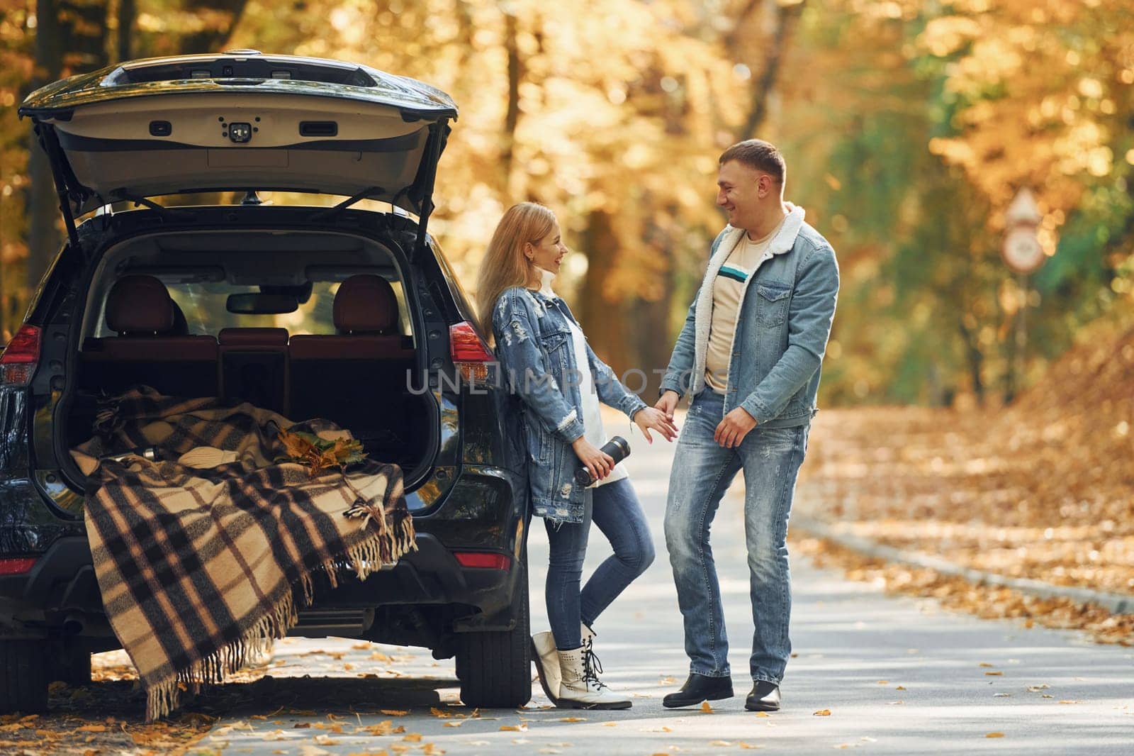 Opened trunk. Couple standing on the road in park near automobile.