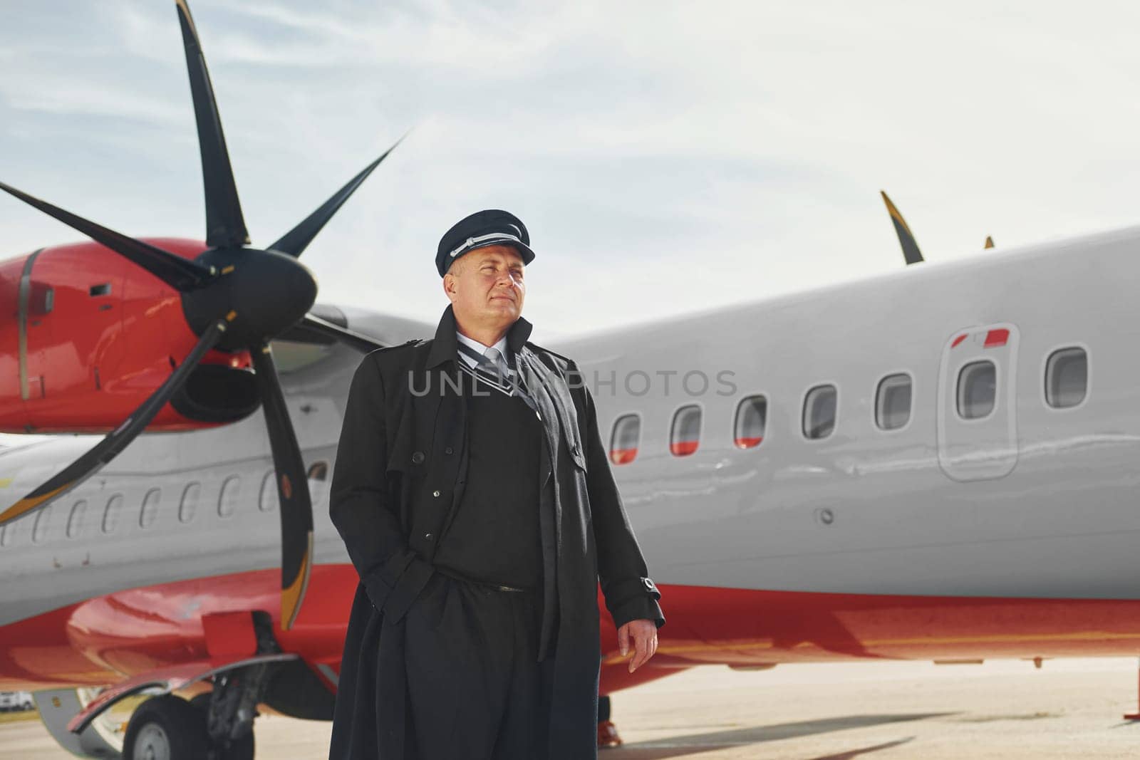 Looking far away. Pilot in formal black uniform is standing outdoors near plane.