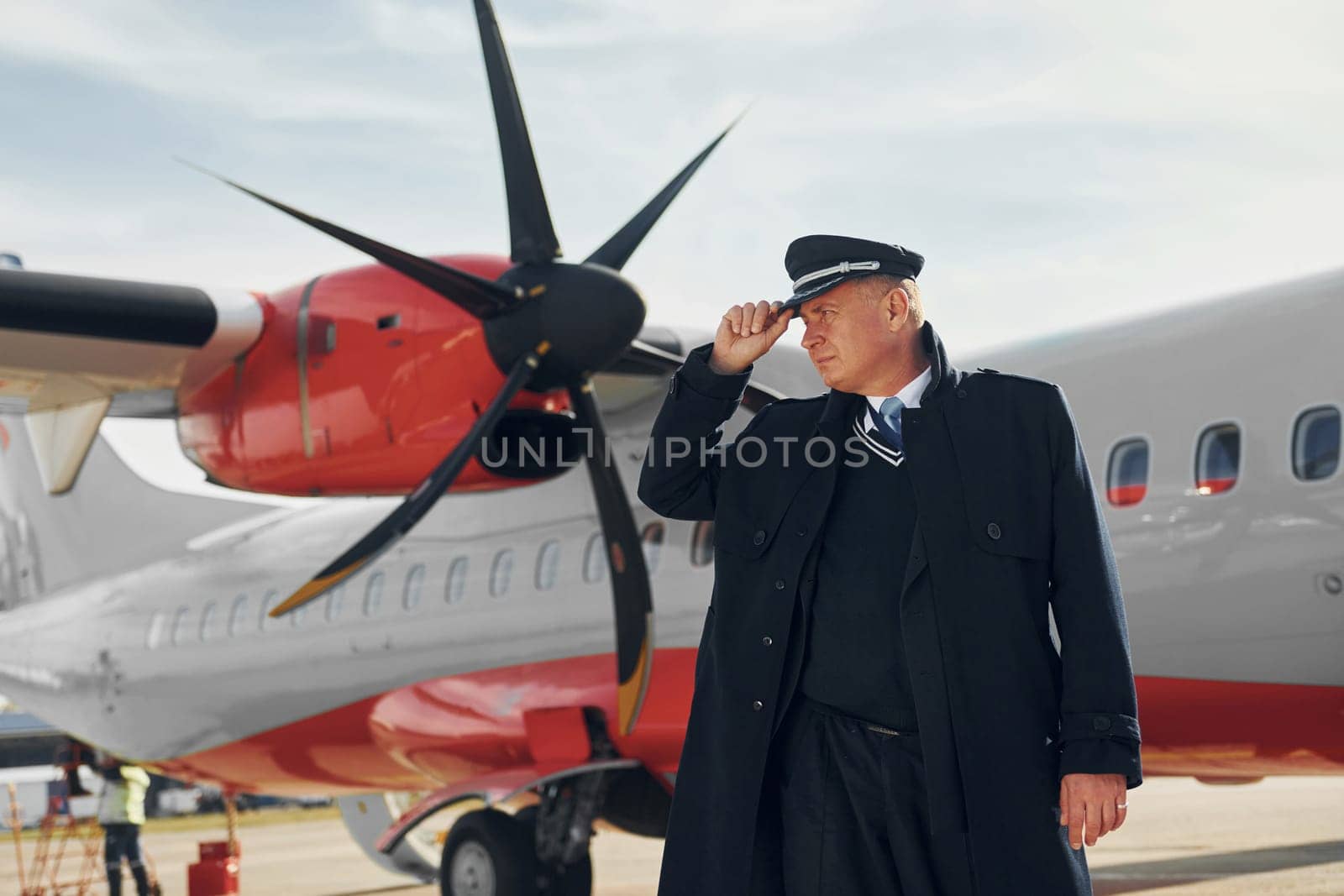 Looking far away. Pilot in formal black uniform is standing outdoors near plane.