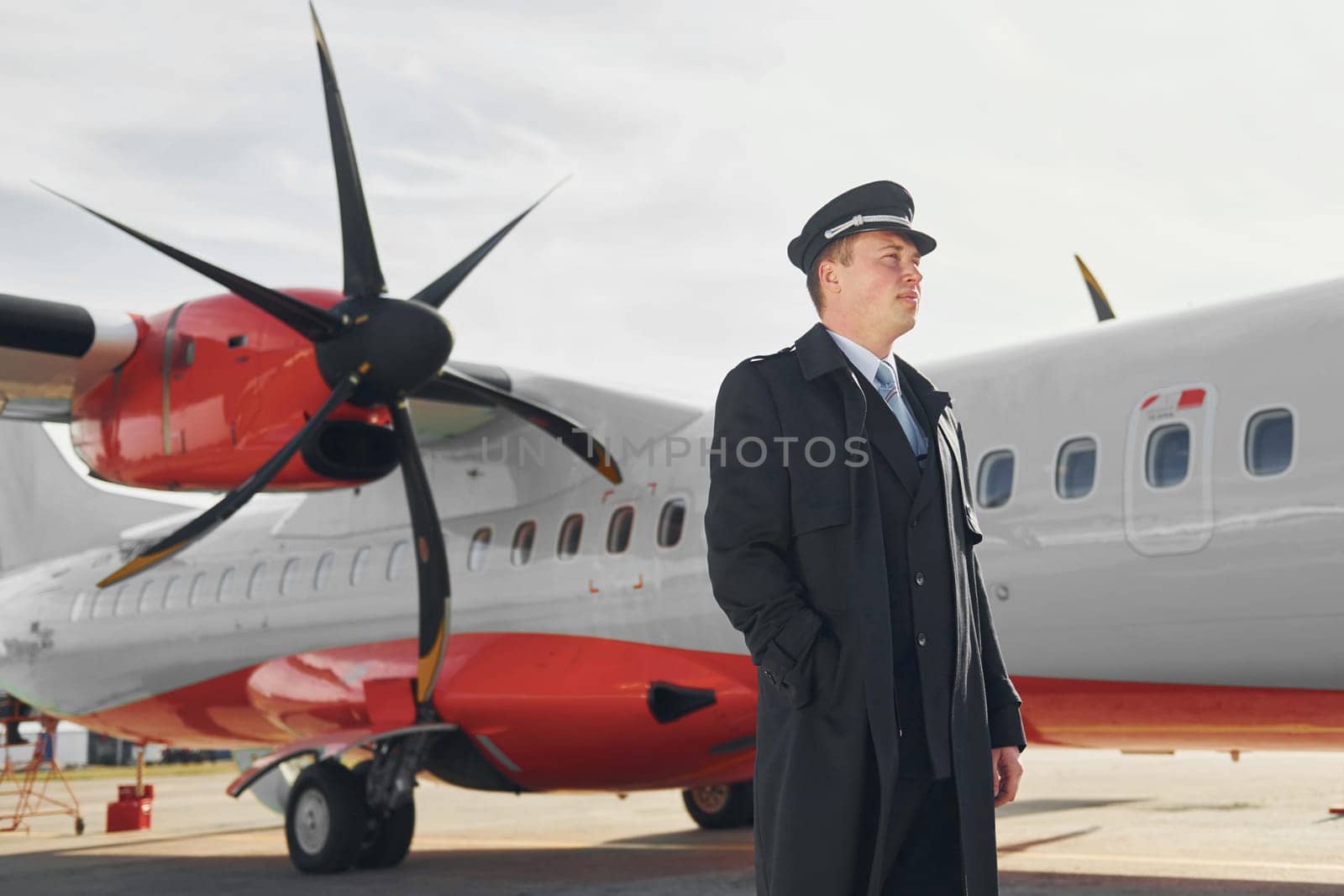Looking far away. Pilot in formal black uniform is standing outdoors near plane.