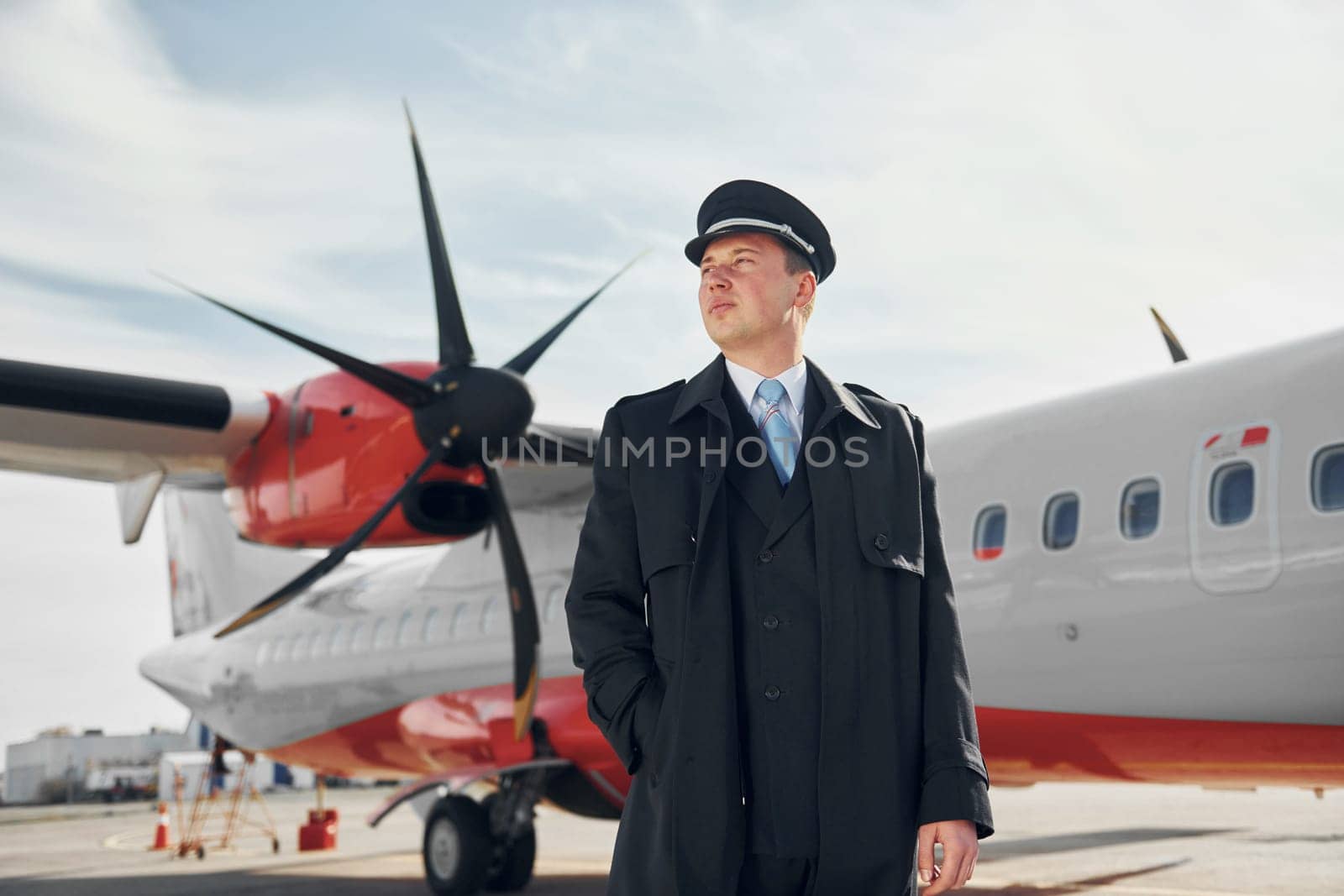 Looking far away. Pilot in formal black uniform is standing outdoors near plane.