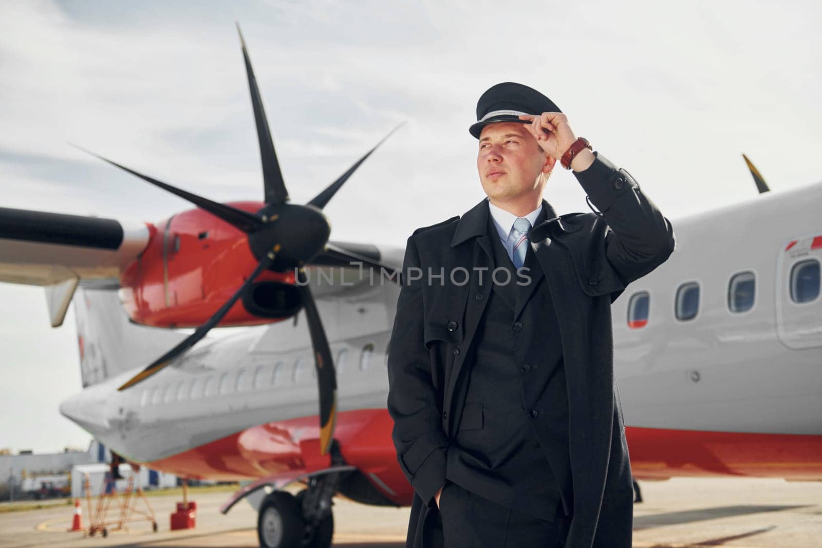 Looking far away. Pilot in formal black uniform is standing outdoors near plane.