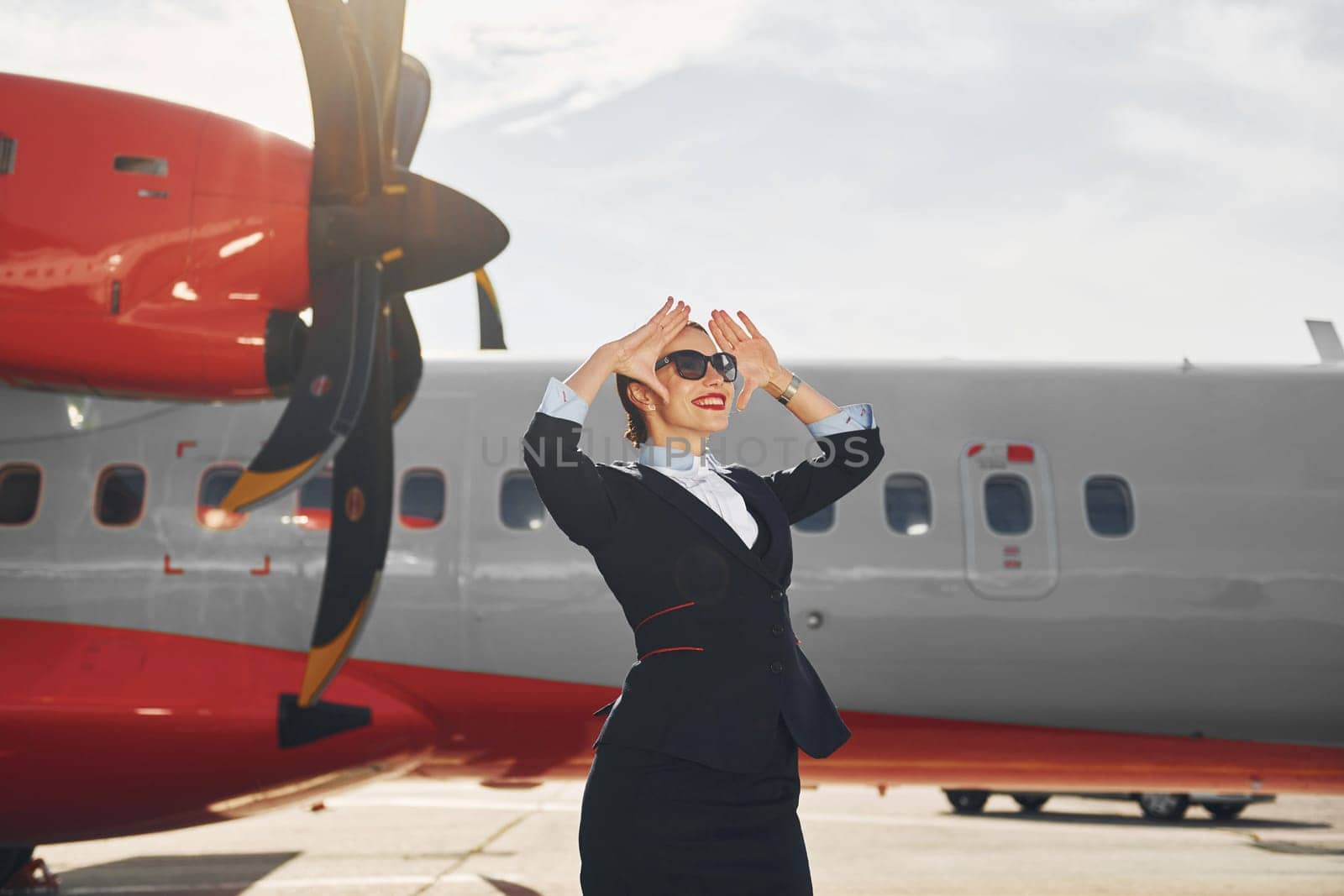 Looking far away. Young stewardess that is in formal black clothes is standing outdoors near plane.