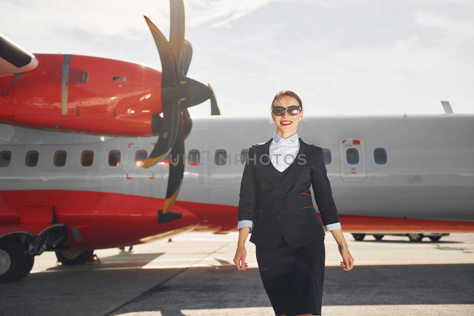 Looking far away. Young stewardess that is in formal black clothes is standing outdoors near plane.