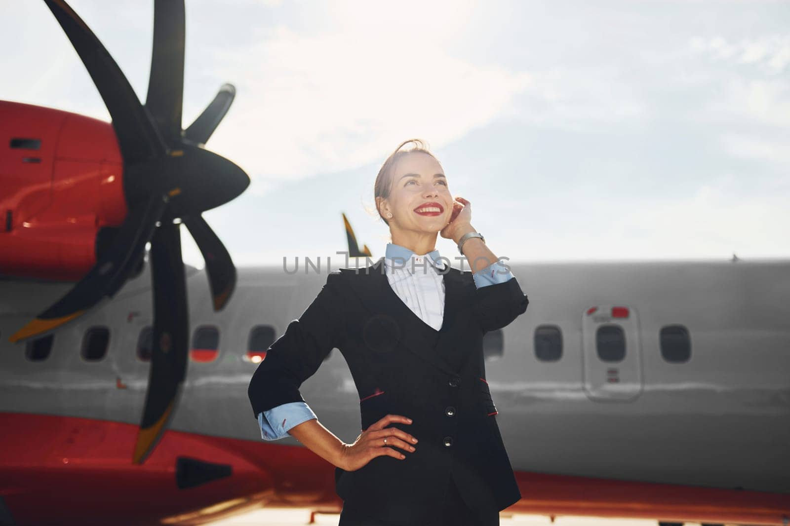 At daytime. Young stewardess that is in formal black clothes is standing outdoors near plane.