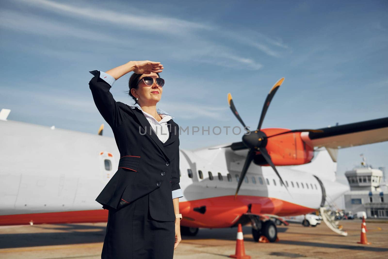 At daytime. Young stewardess that is in formal black clothes is standing outdoors near plane.