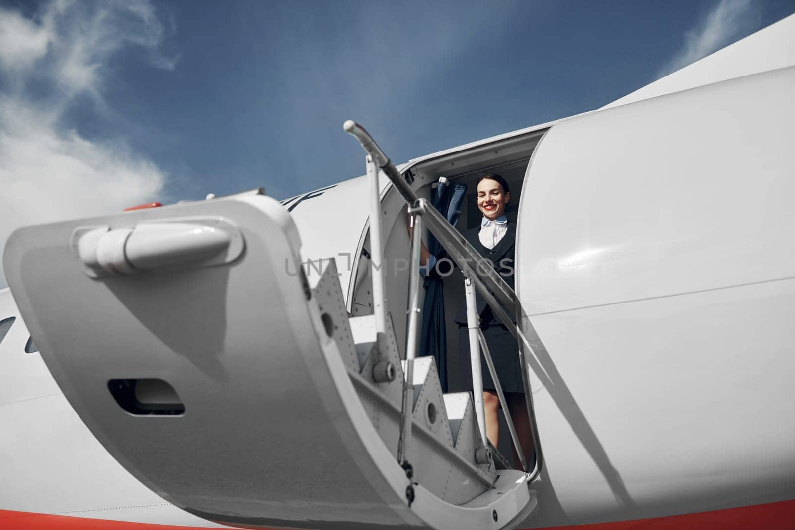 At daytime. Young stewardess that is in formal black clothes is standing outdoors near plane.