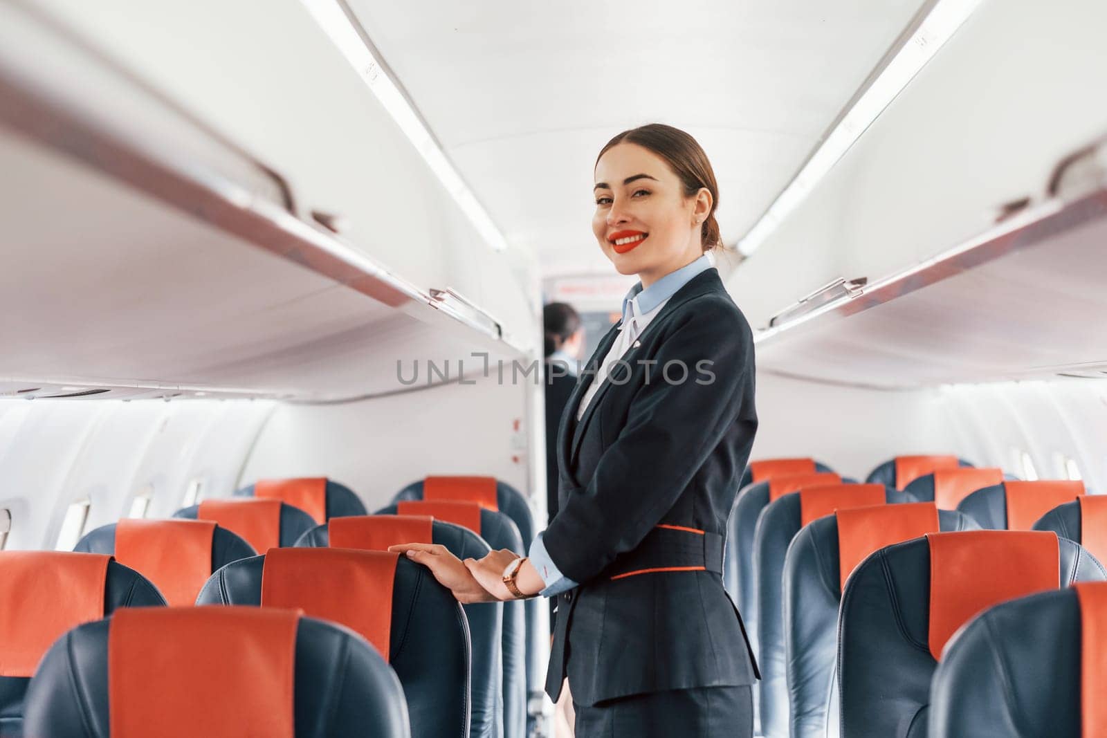 Young stewardess that is in formal black clothes is standing indoors in the plane.