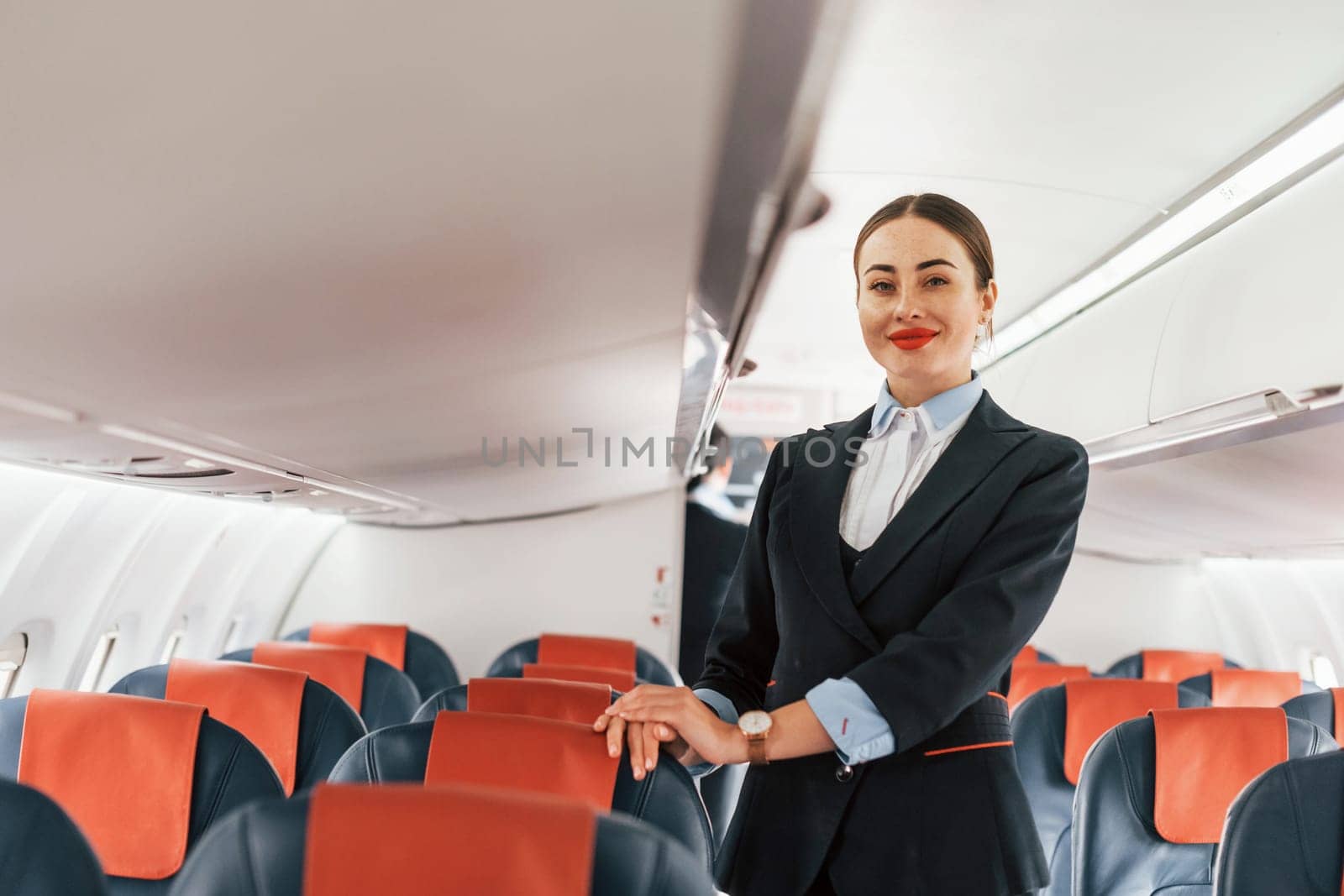 Young stewardess that is in formal black clothes is standing indoors in the plane.