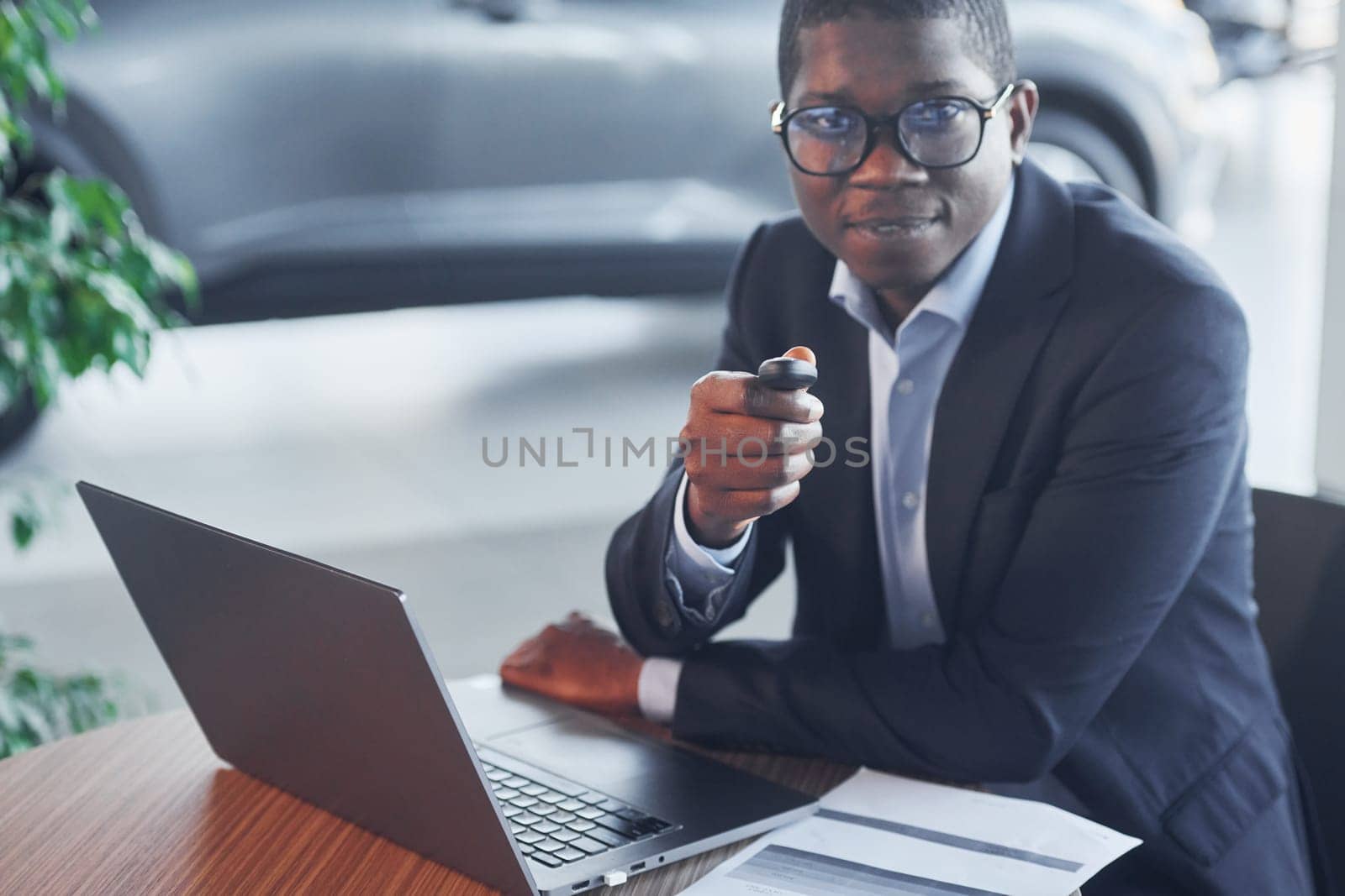 Office worker is sitting in autosalon by table with laptop.