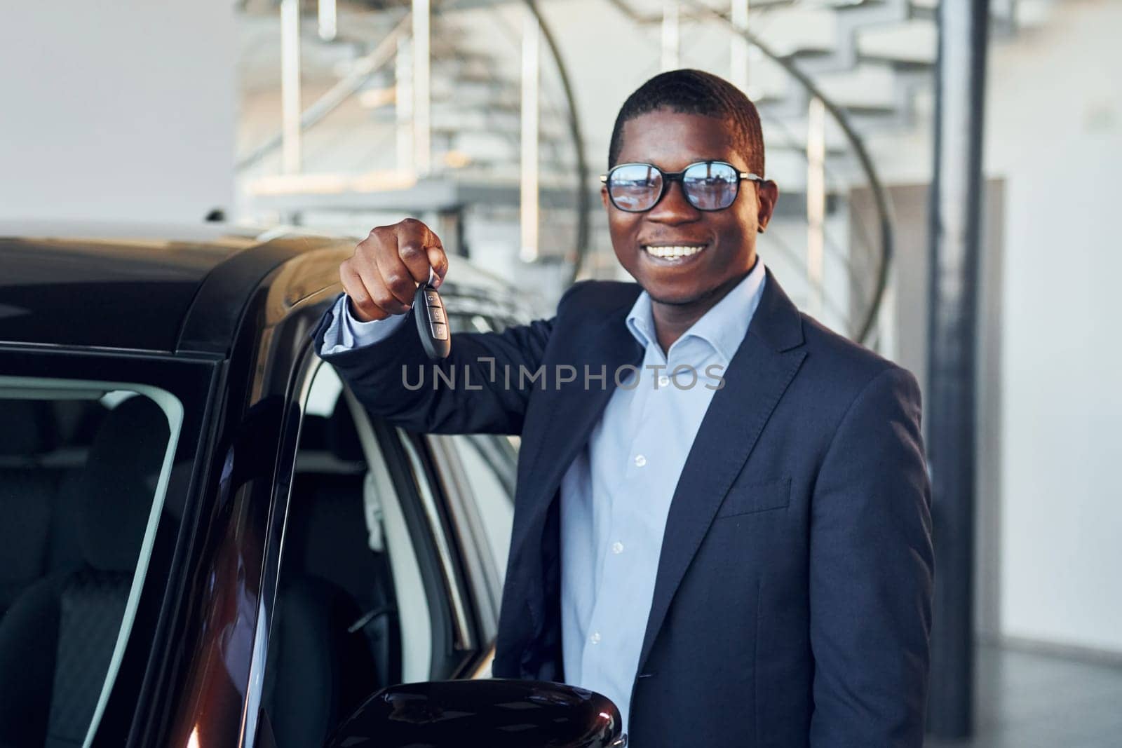 Standing near car. Young african american businessman in black suit is the autosalon.