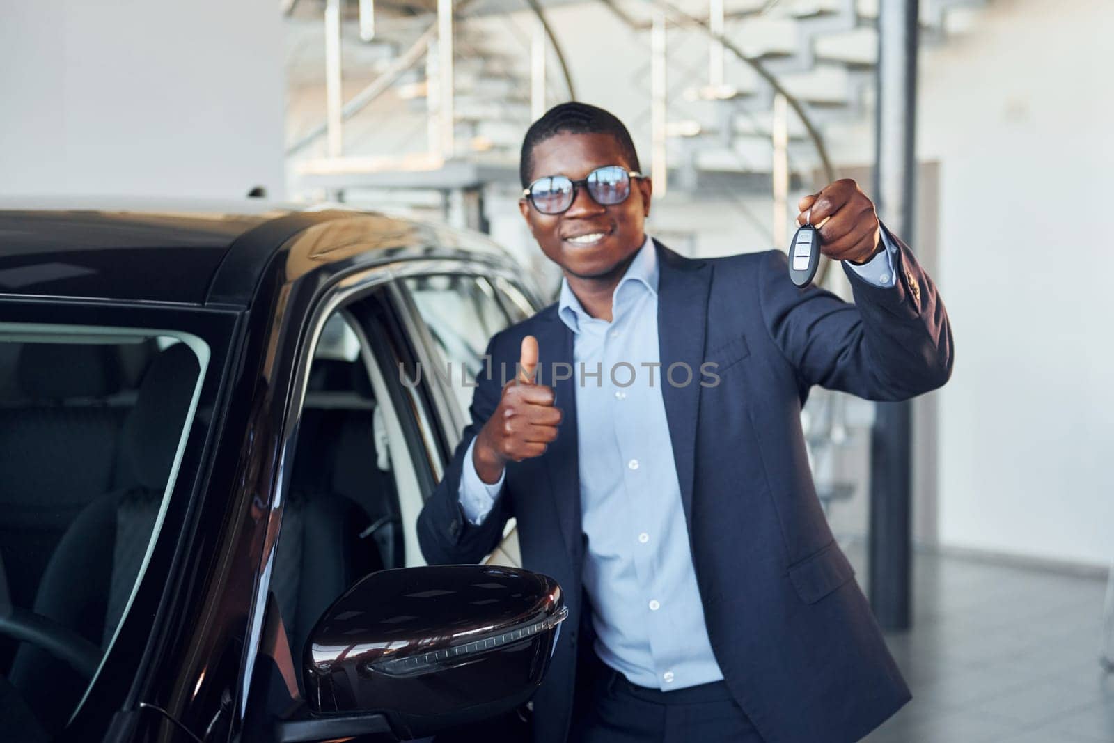 Holding keys in hands. Young african american businessman in black suit is the autosalon.
