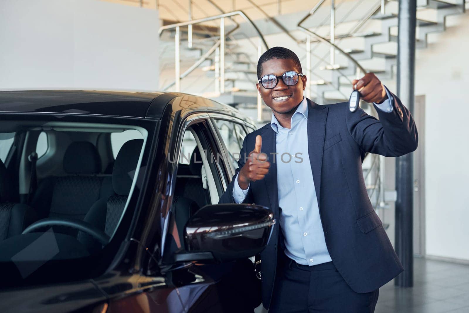 Holding keys in hands. Young african american businessman in black suit is the autosalon.
