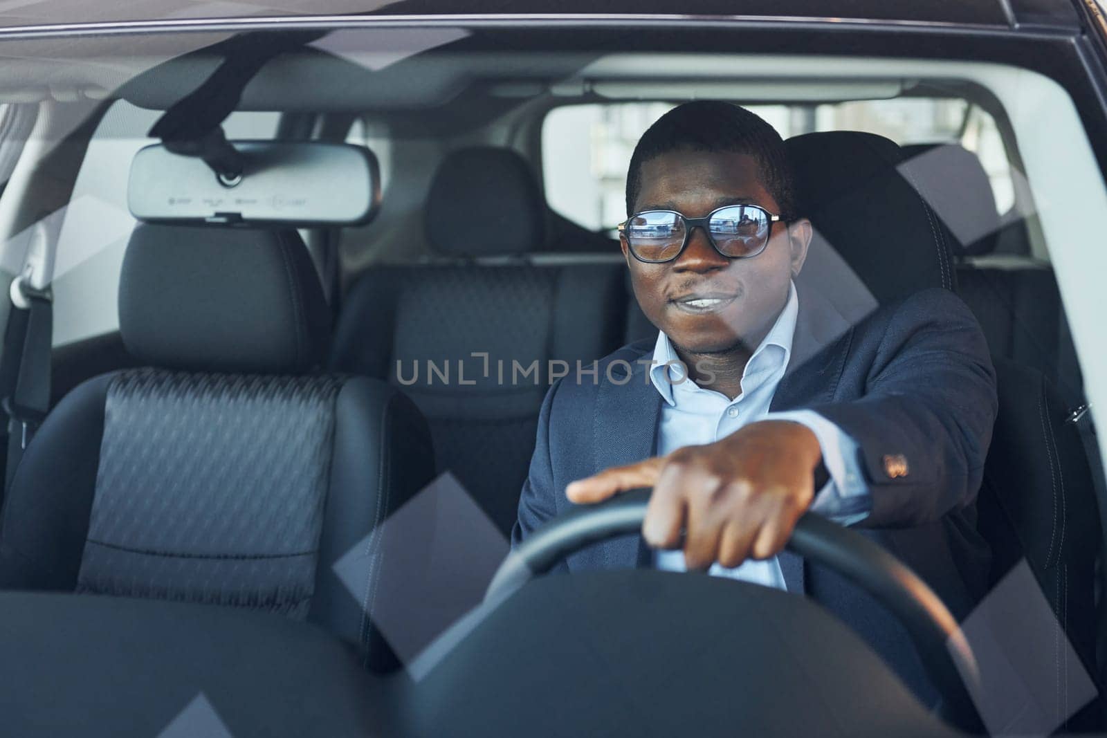 Positive mood. Young african american businessman in black suit is in the automobile.