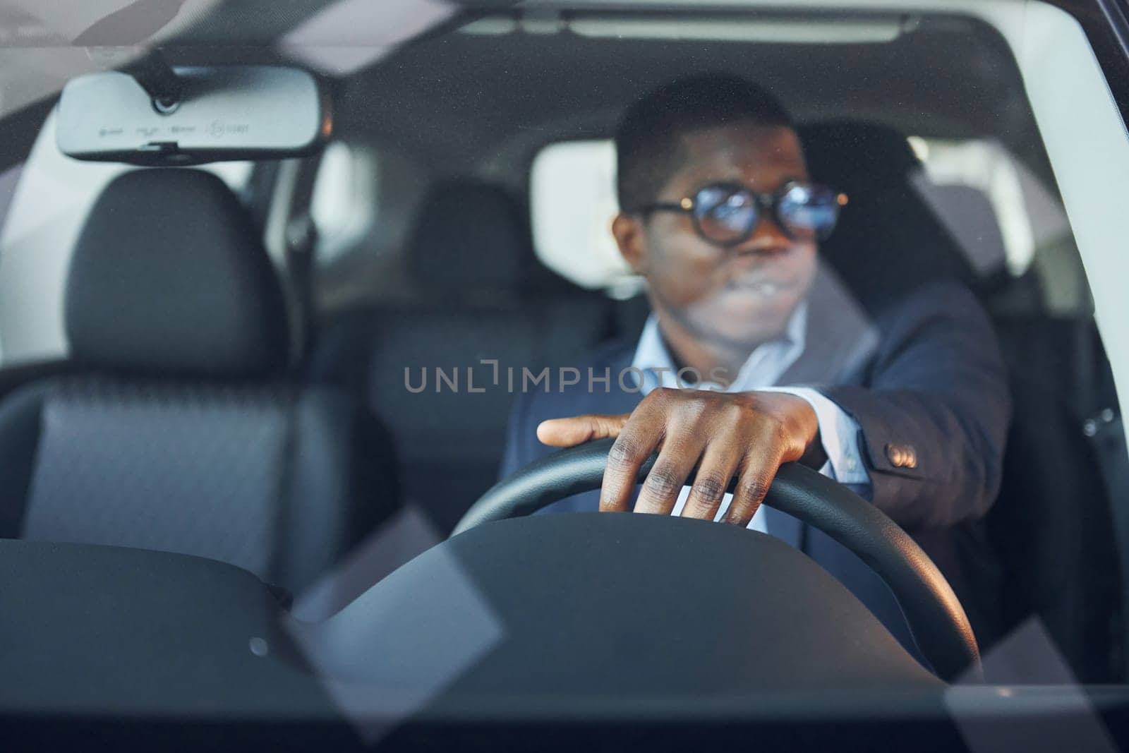 Front view. Young african american businessman in black suit is in the automobile.