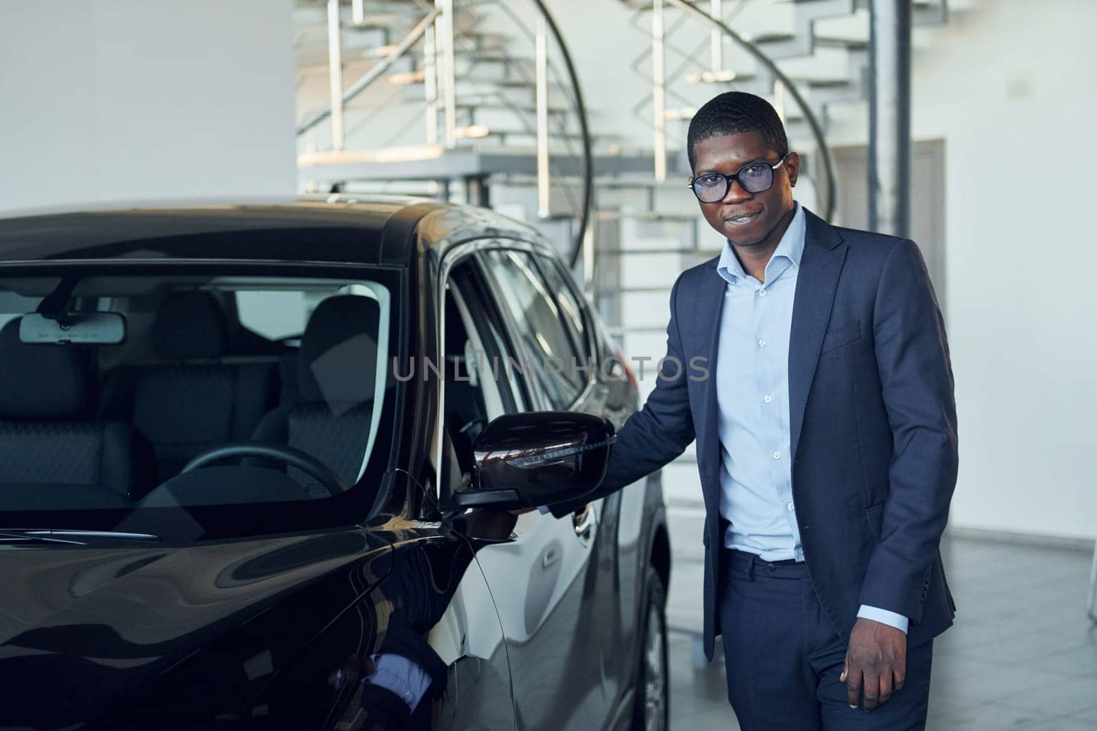Looking at car. Young african american businessman in black suit is the autosalon.