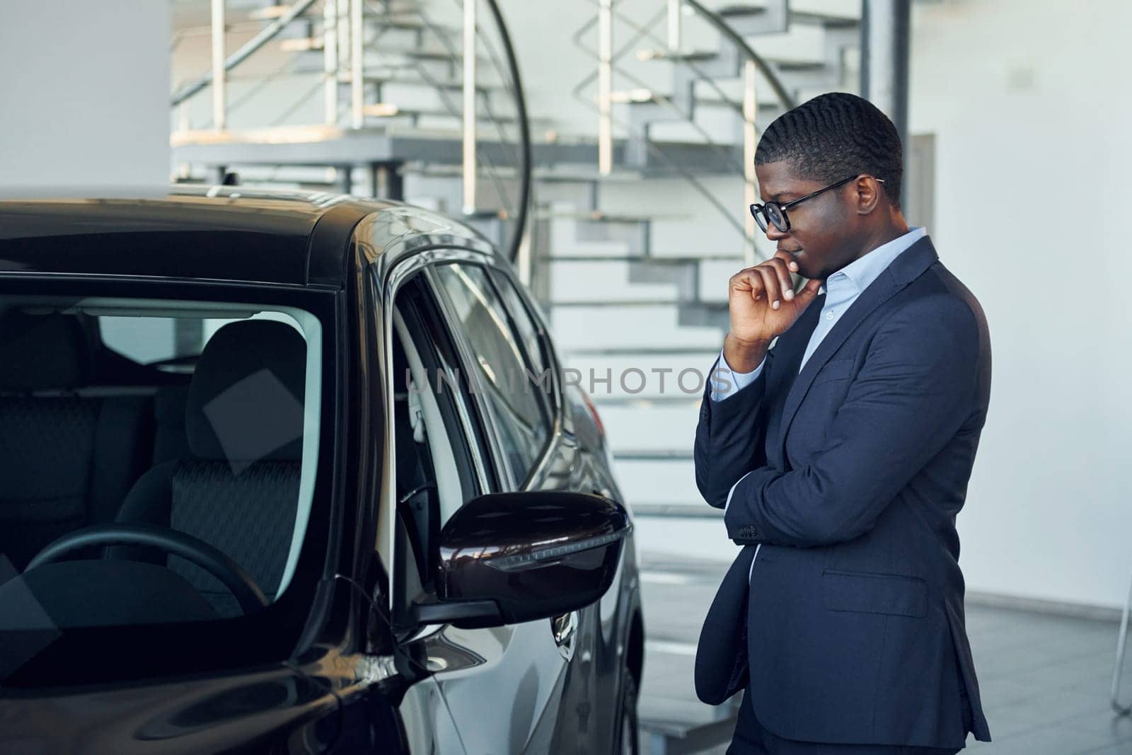 Looking at car. Young african american businessman in black suit is the autosalon.