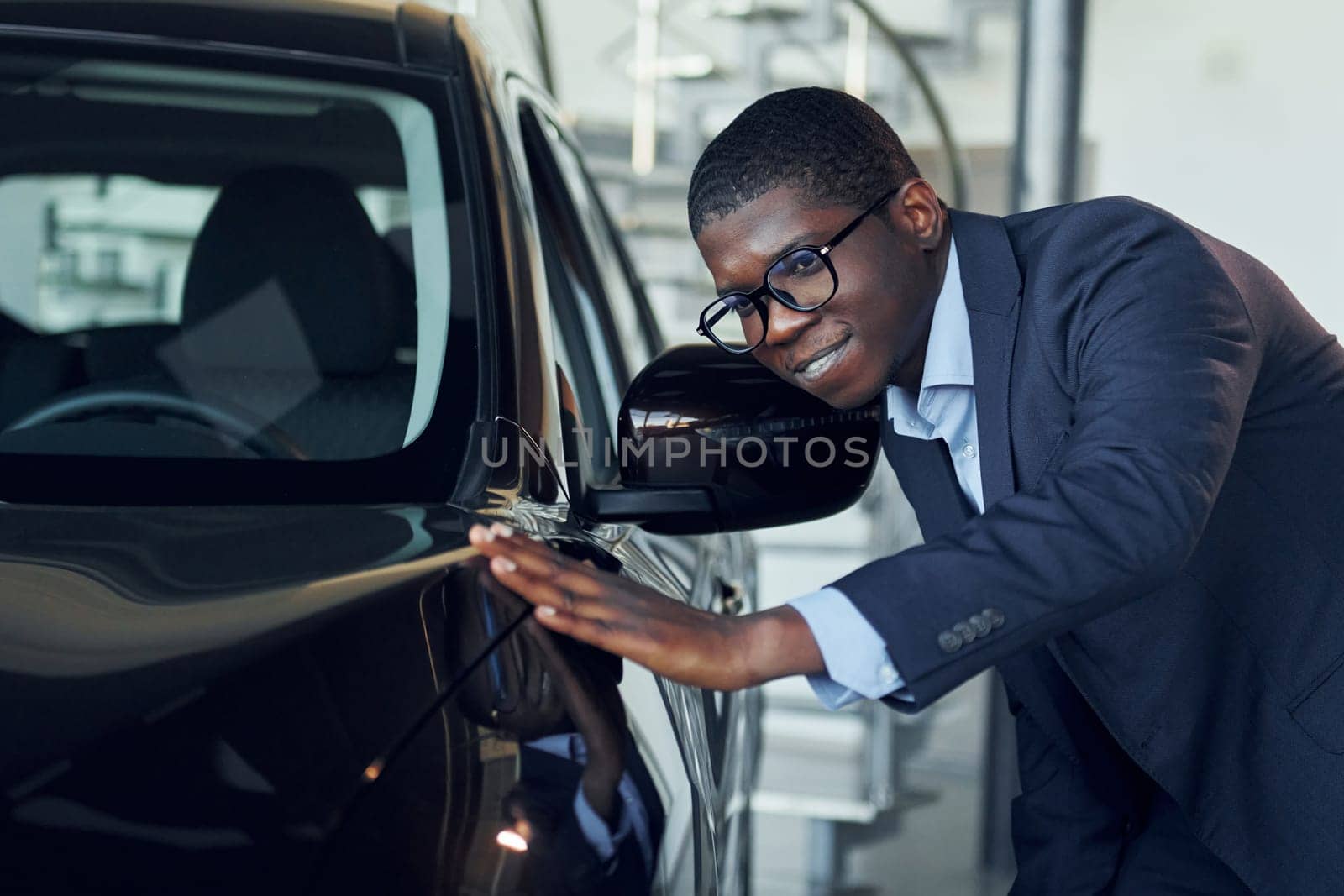 Looking at car. Young african american businessman in black suit is the autosalon.