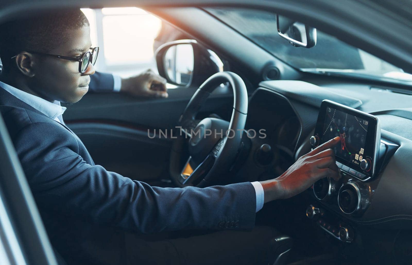 Using front control panel. Young african american businessman in black suit is in the automobile.