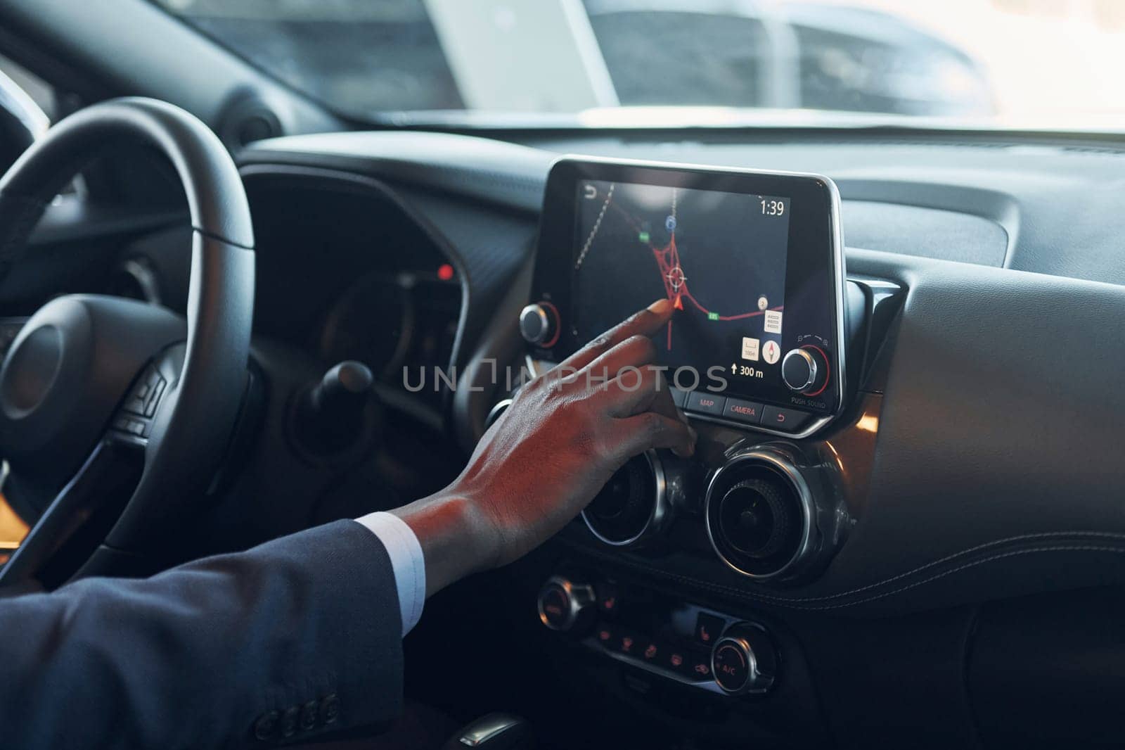 Using front control panel. Young african american businessman in black suit is in the automobile.