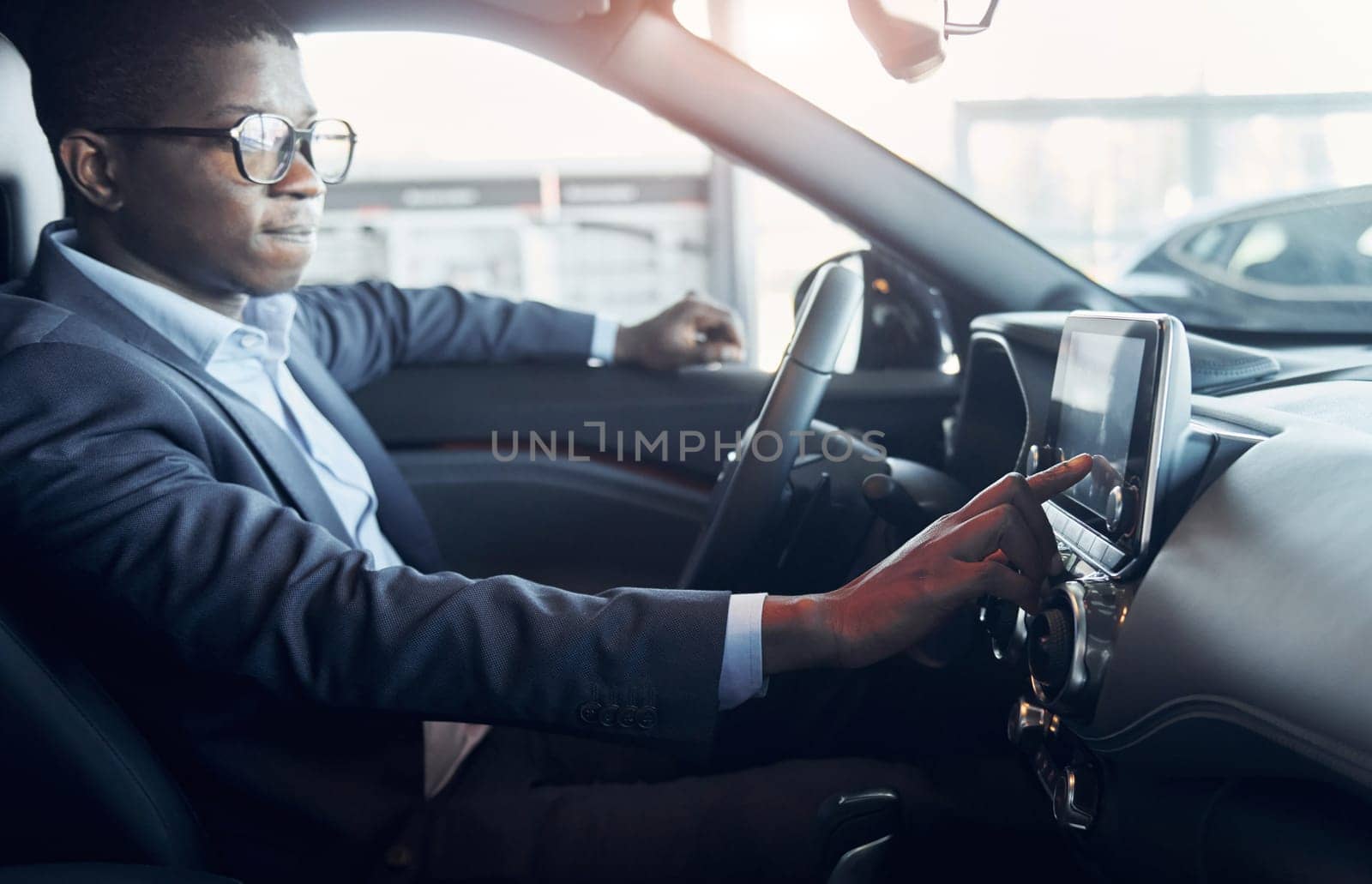 Using front control panel. Young african american businessman in black suit is in the automobile.