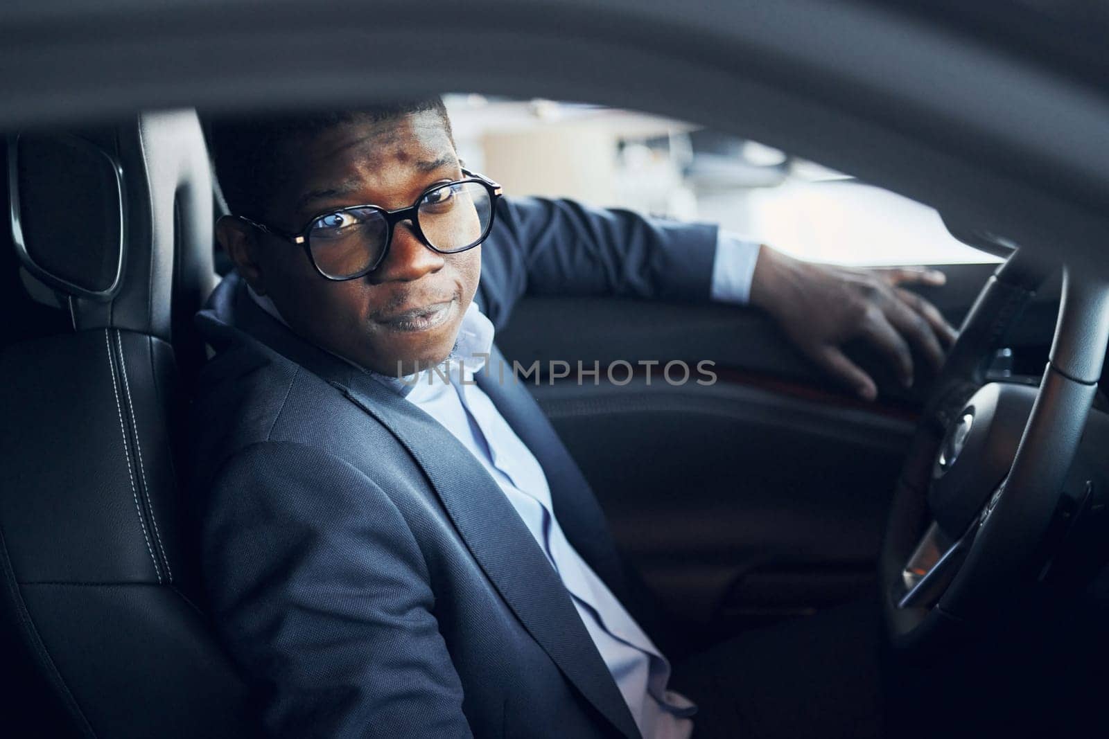 In glasses. Young african american businessman in black suit is in the automobile.