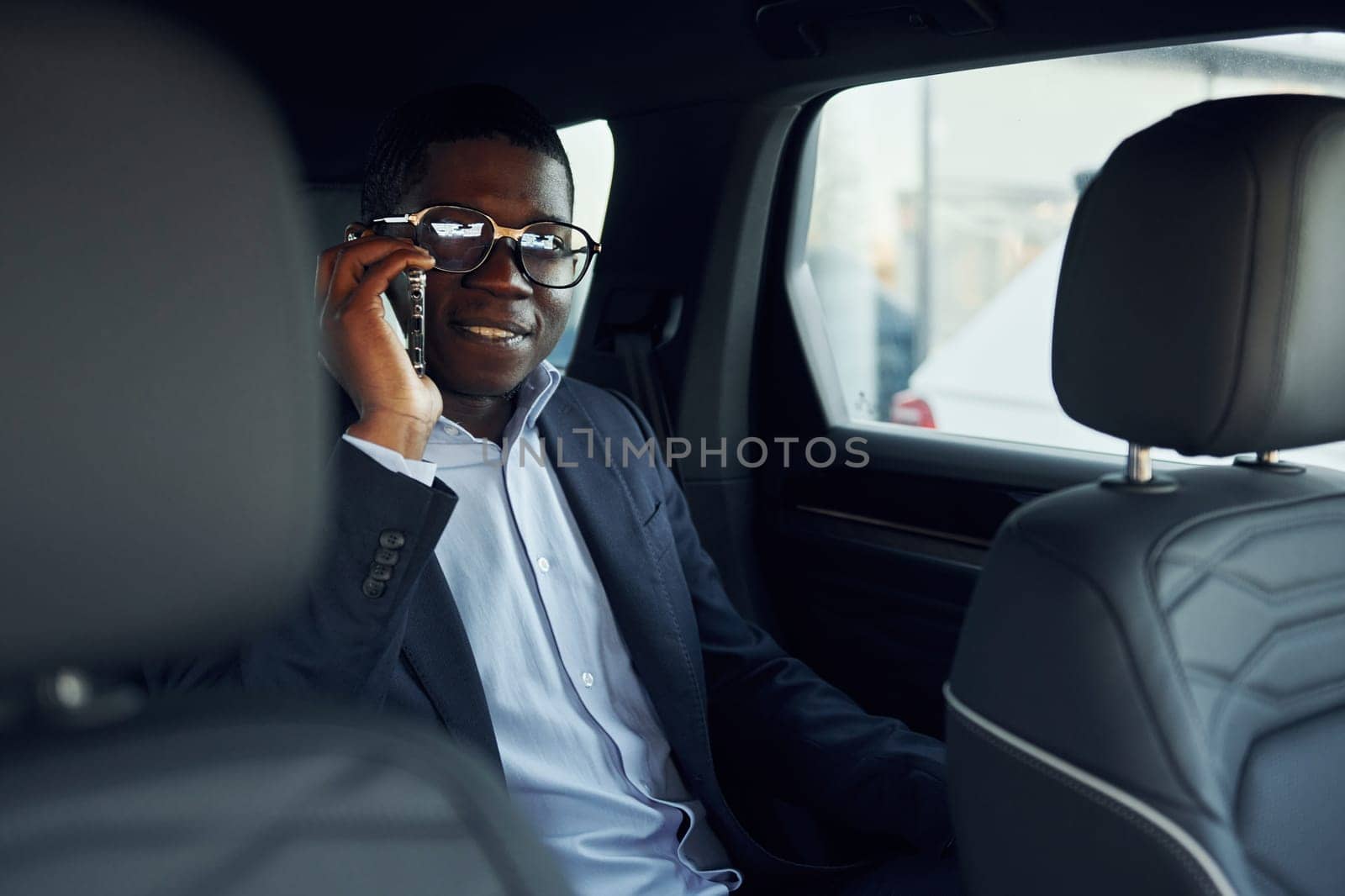 In glasses. Young african american businessman in black suit is in the automobile.