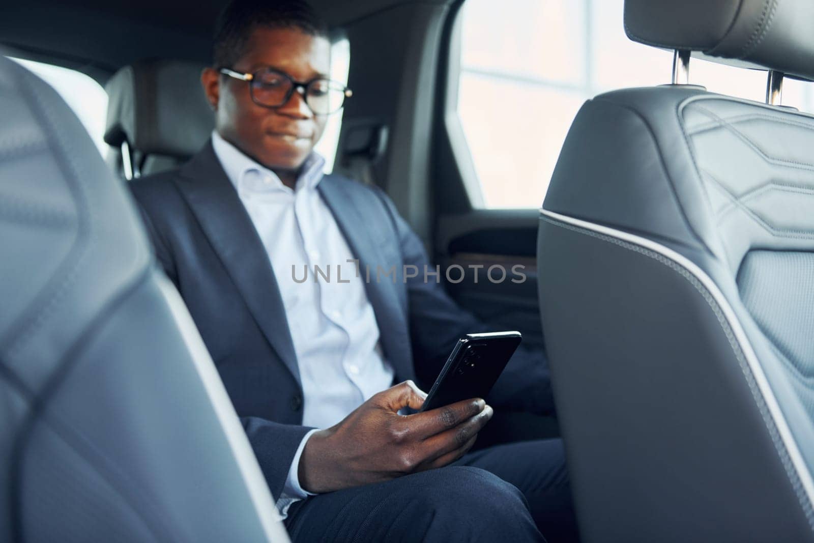 In glasses. Young african american businessman in black suit is in the automobile.