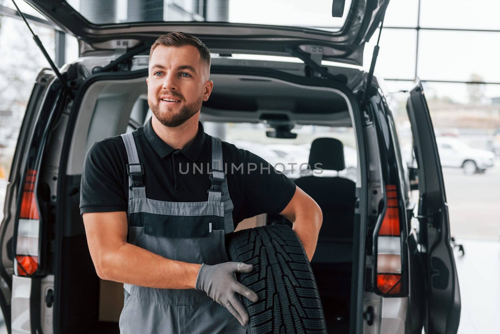 Holding tire. Man in uniform is working in the autosalon at daytime.
