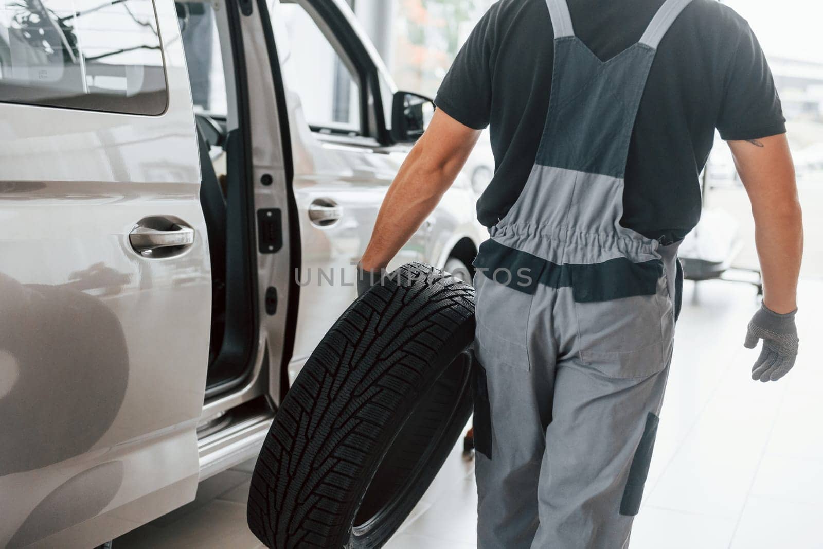With tire in hands. Man in uniform is working in the autosalon at daytime.