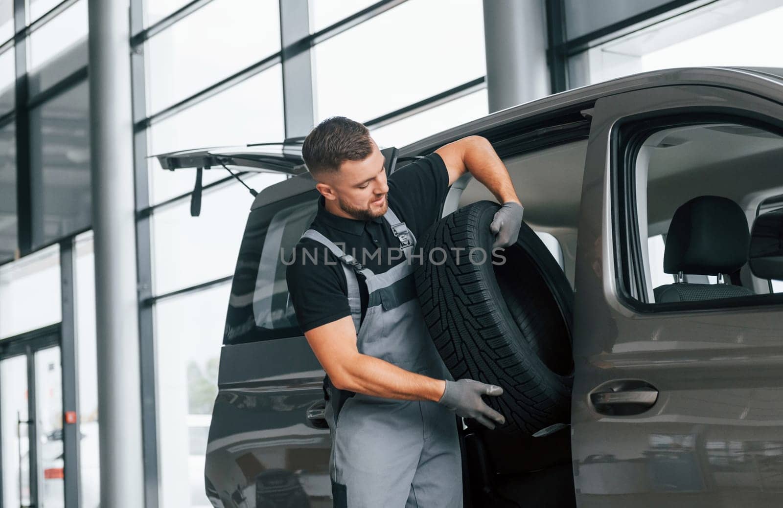 With tire in hands. Man in uniform is working in the autosalon at daytime.