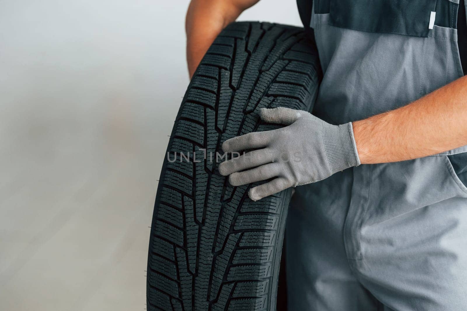 Man in uniform is working in the autosalon at daytime. Holding tire in hands.