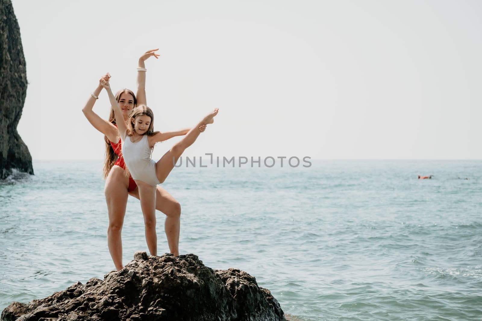 Woman and her daughter practicing balancing yoga pose on one leg up together on rock in the sea. Silhouette mother and daughter doing yoga at beach by panophotograph