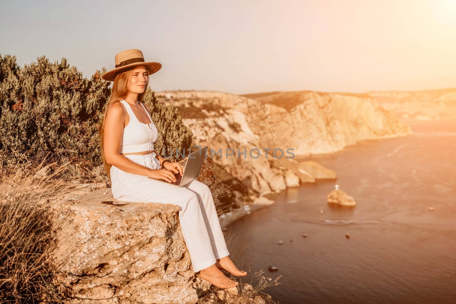 Successful business woman in yellow hat working on laptop by the sea. Pretty lady typing on computer at summer day outdoors. Freelance, travel and holidays concept.