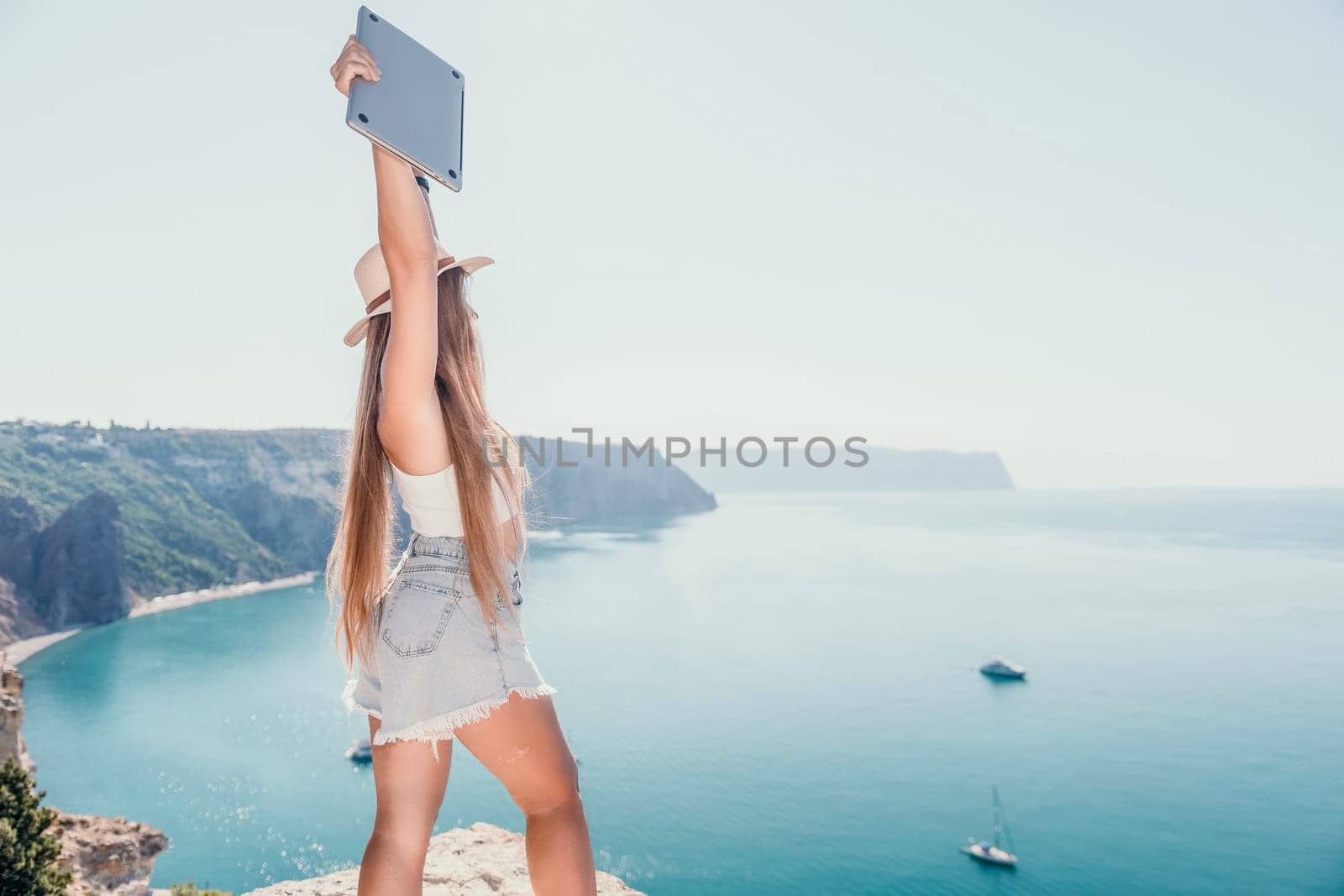 Woman sea laptop. Back view of happy young woman, female freelancer in straw hat working on laptop, keeping arms raised and cellebrating success while standing over blue sea beach. Work from anywhere. by panophotograph