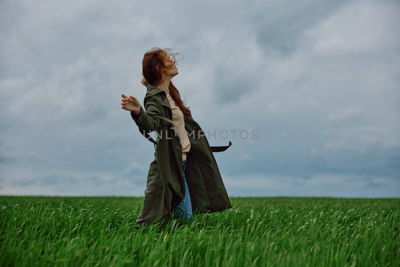 a woman in a dark coat with long red hair stands in a field against a cloudy sky in windy weather by Vichizh