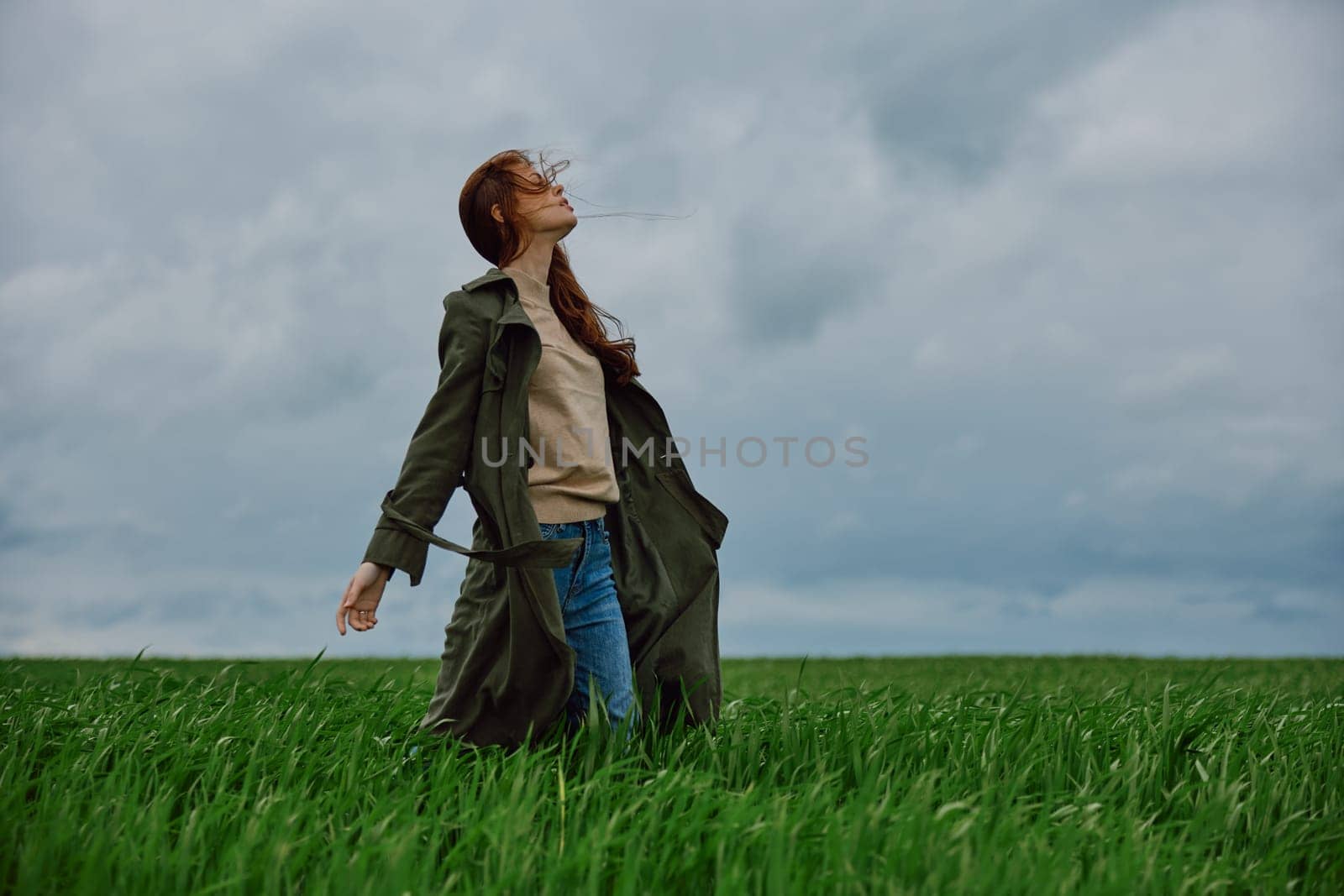 a woman in a dark coat with long red hair stands in a field against a cloudy sky in windy weather by Vichizh