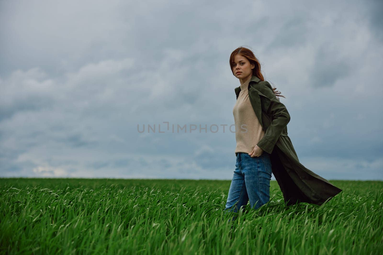 a woman in a coat stands in a green field in cloudy, windy weather holding her hands in her pockets. High quality photo