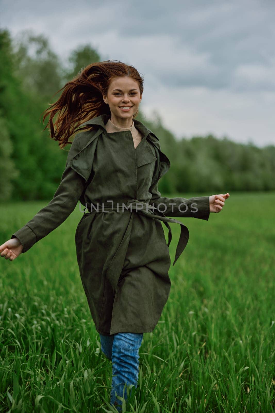 a beautiful woman in a long raincoat runs across a field in high grass in spring in cloudy weather by Vichizh