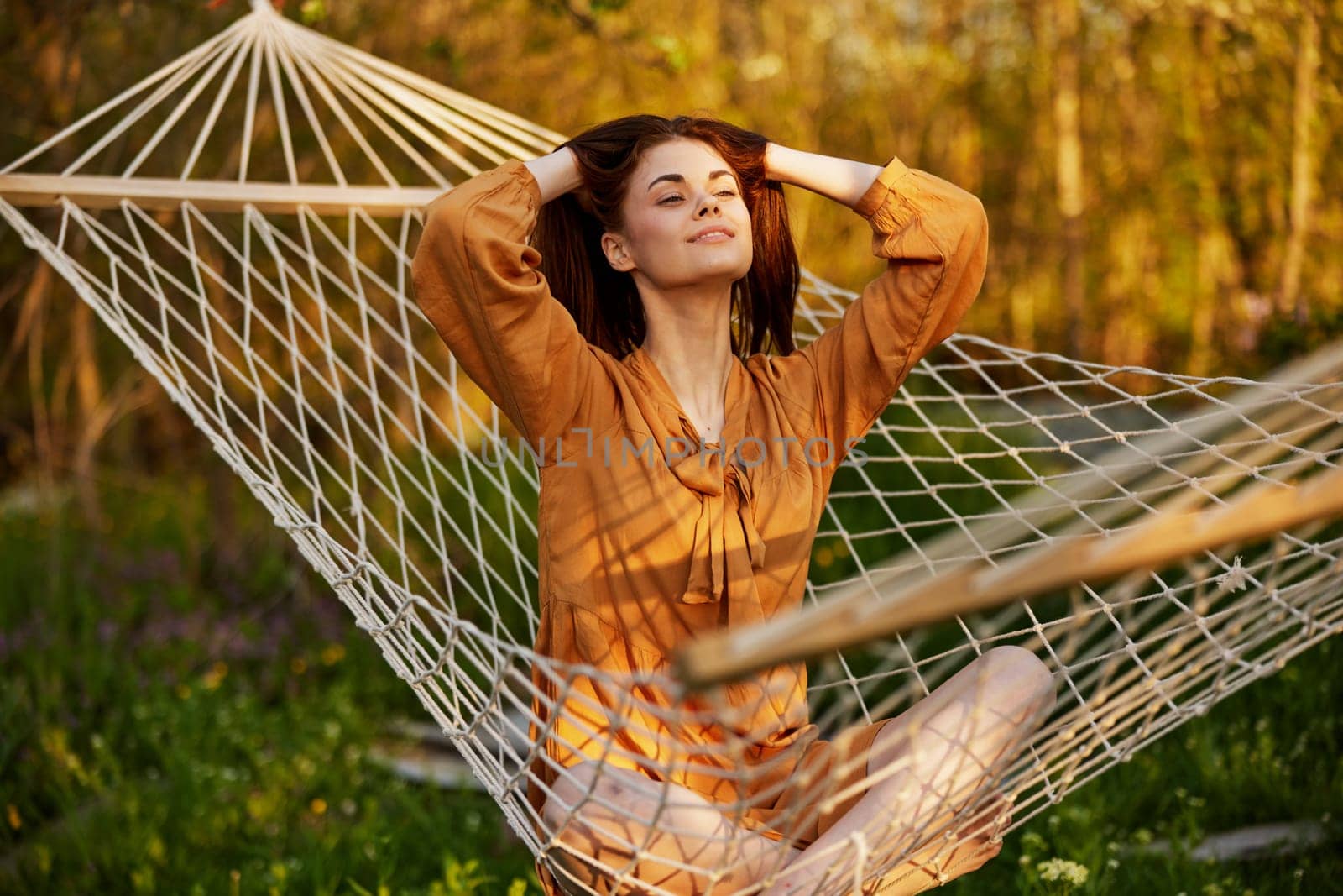 a happy woman in a long orange dress is resting lying in a hammock at the dacha, straightening her long hair and looking away, illuminated by the summer sun during sunset. High quality photo