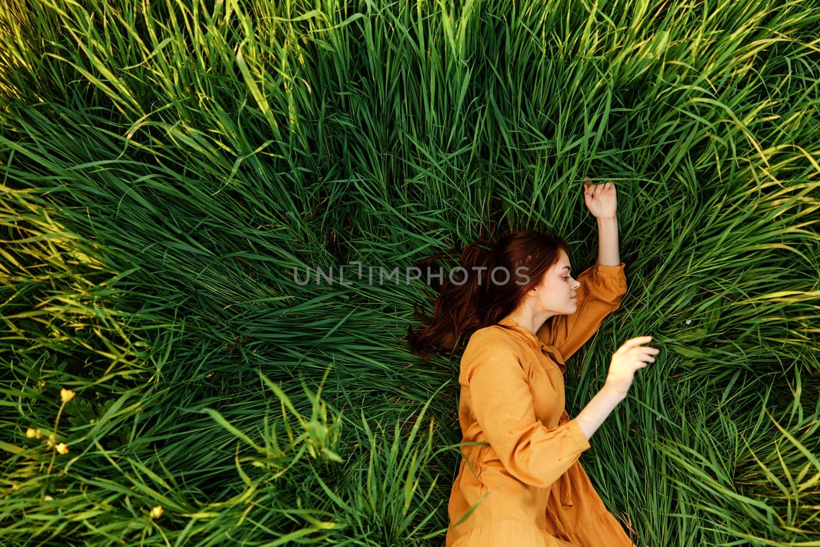 an elegant woman lies in the tall green grass in a long summer orange dress and relaxed posing with her hands enjoying nature and sunny weather with her eyes closed. High quality photo