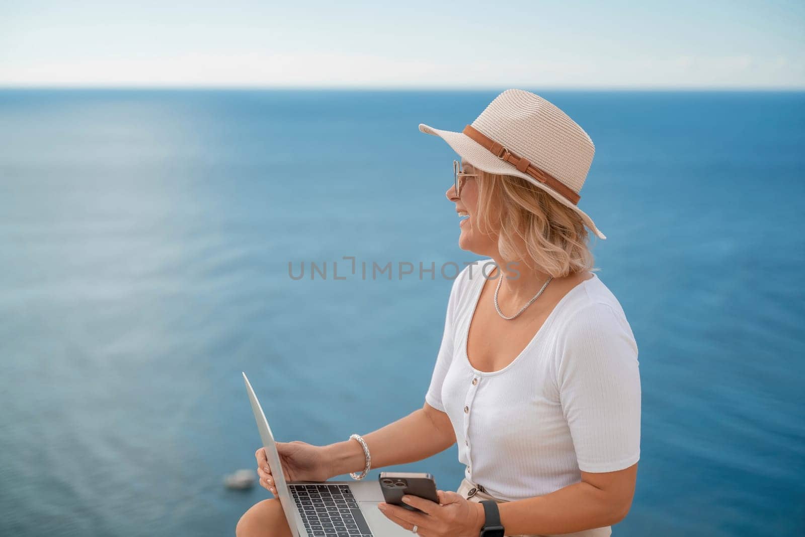 Freelance women sea working on the computer. Good looking middle aged woman typing on a laptop keyboard outdoors with a beautiful sea view. The concept of remote work