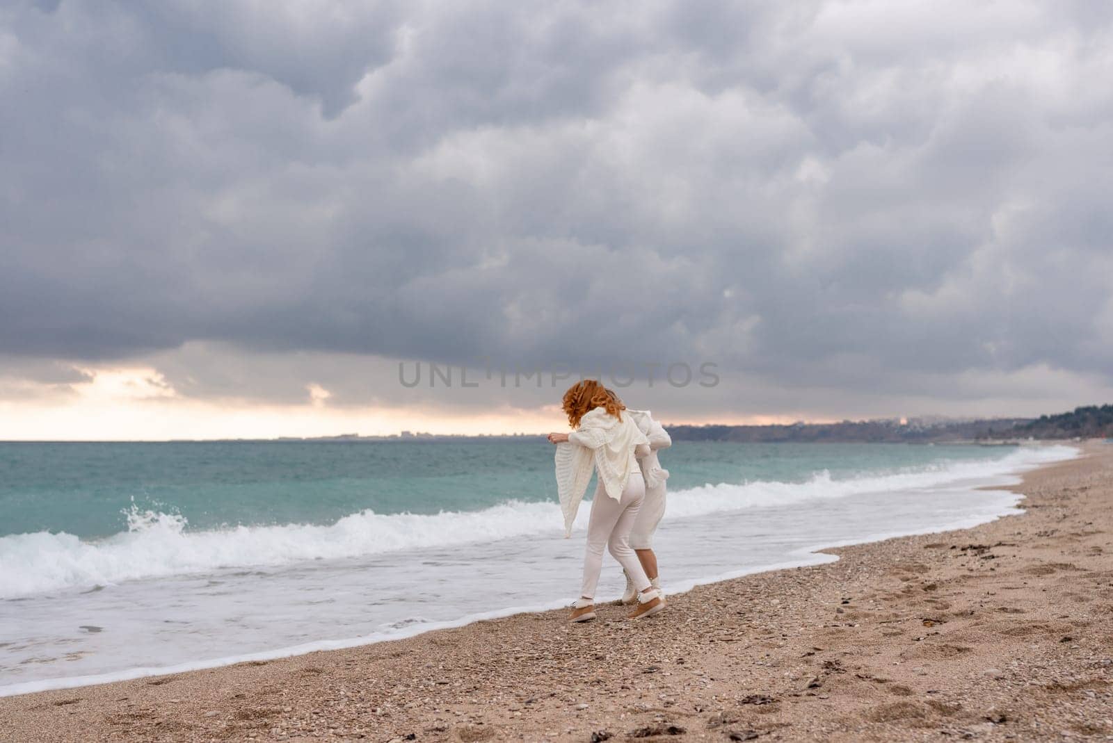 Women sea walk friendship spring. Two girlfriends, redhead and blonde, middle-aged walk along the sandy beach of the sea, dressed in white clothes. Against the backdrop of a cloudy sky and the winter sea. Weekend concept. by Matiunina