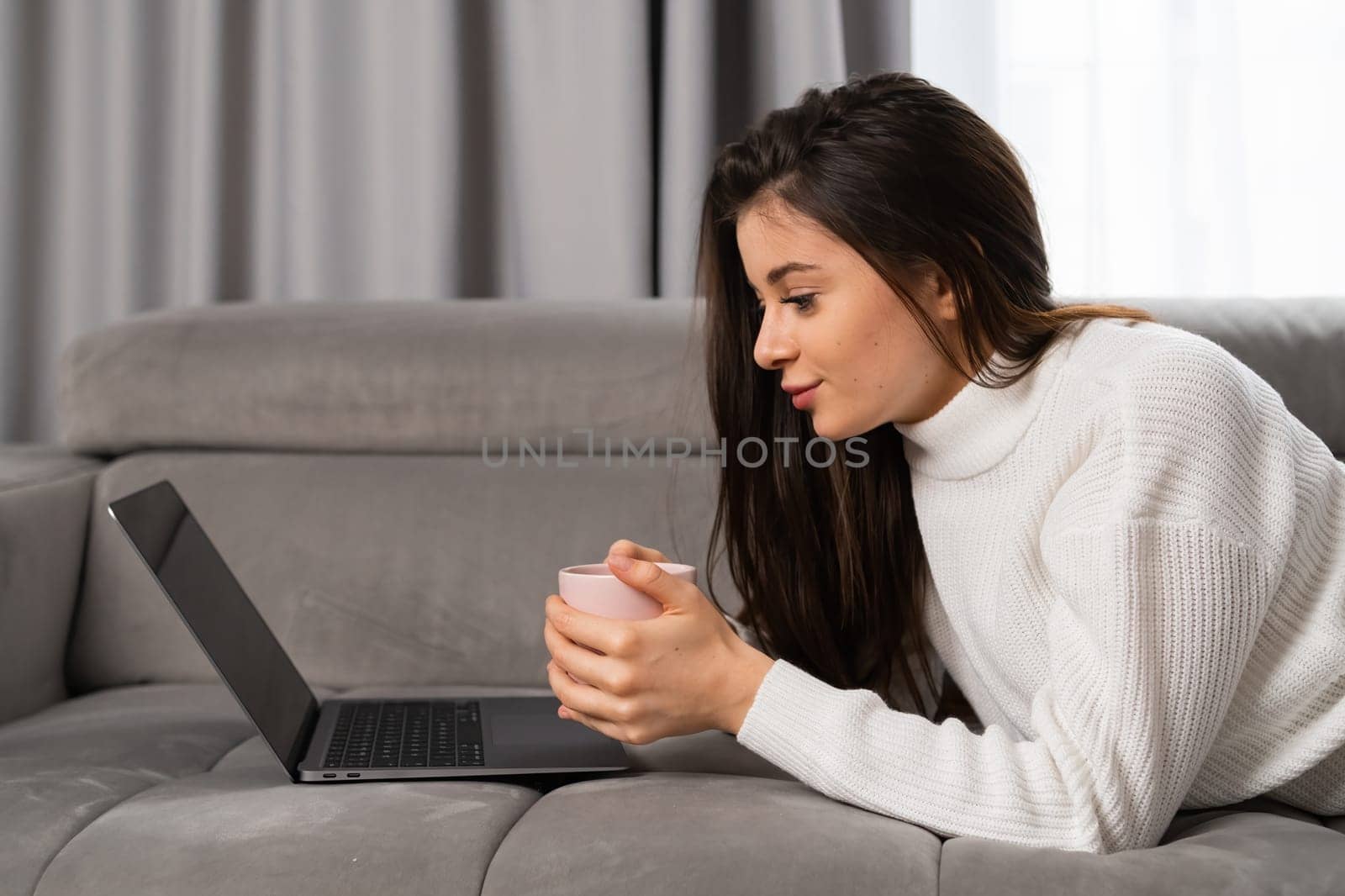 Young beautiful woman relaxing, holding a cup, watching movie using a laptop, laying on the sofa