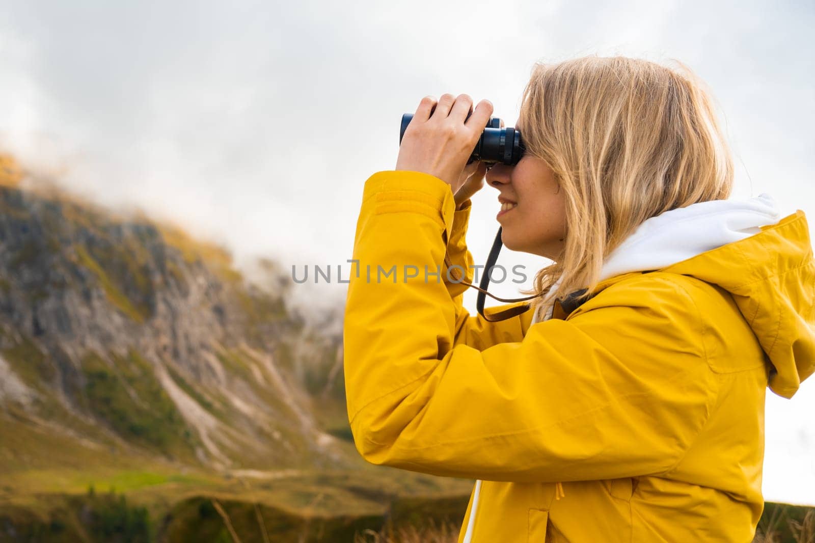 Side view of traveling girl looking through binoculars in mountains covered by clouds in a sunny day