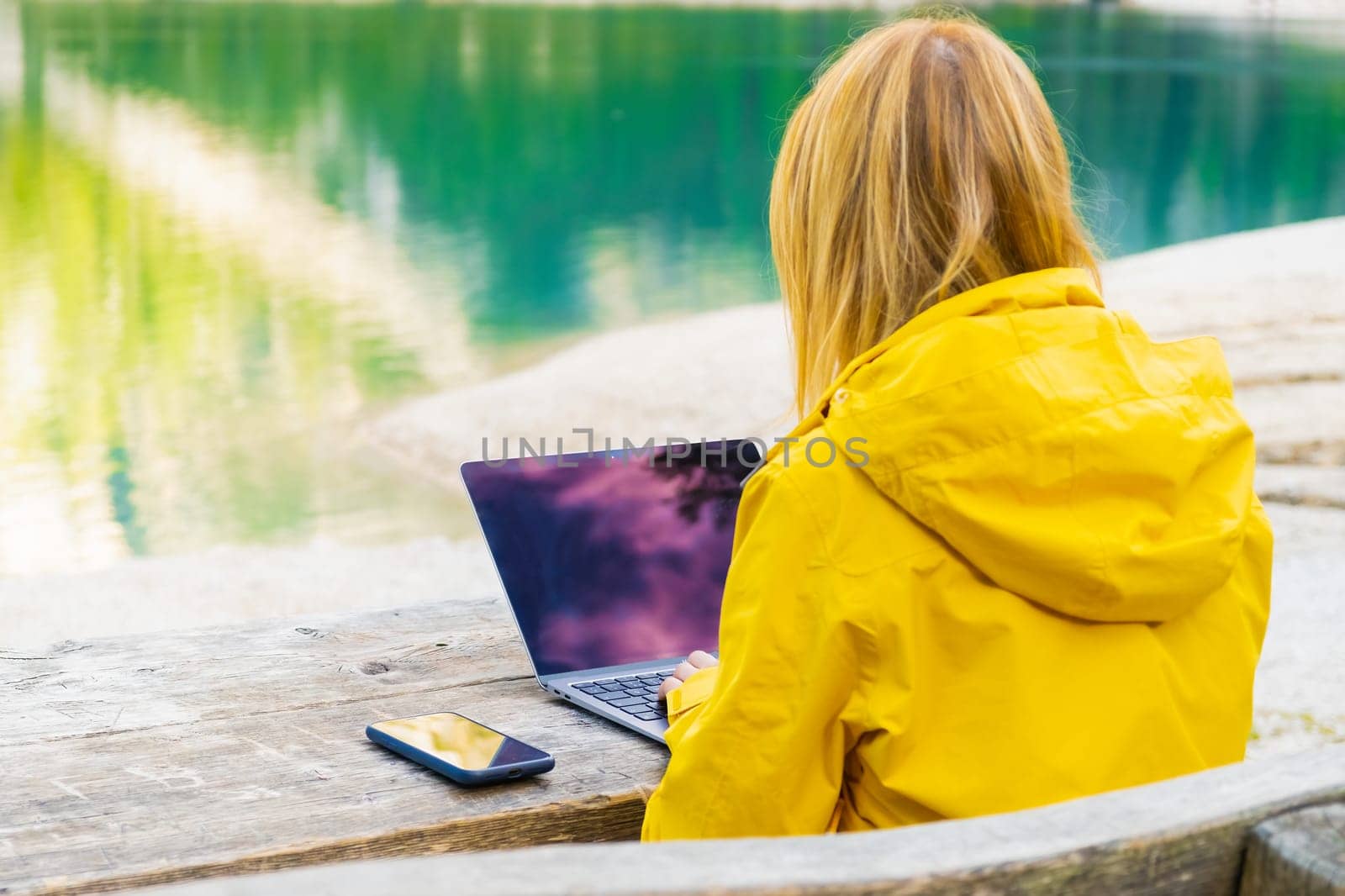 The girl is focused on working at a computer in nature with a beautiful view of the mountains and the lake. Freelancer works remotely from the office in sunny day.