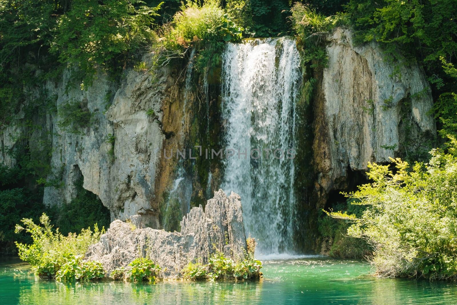 Waterfall flows down from rocky cliff into clear turquoise water at sunlight. Breathtaking view of aquamarine cascade in national park on Plitvice lakes